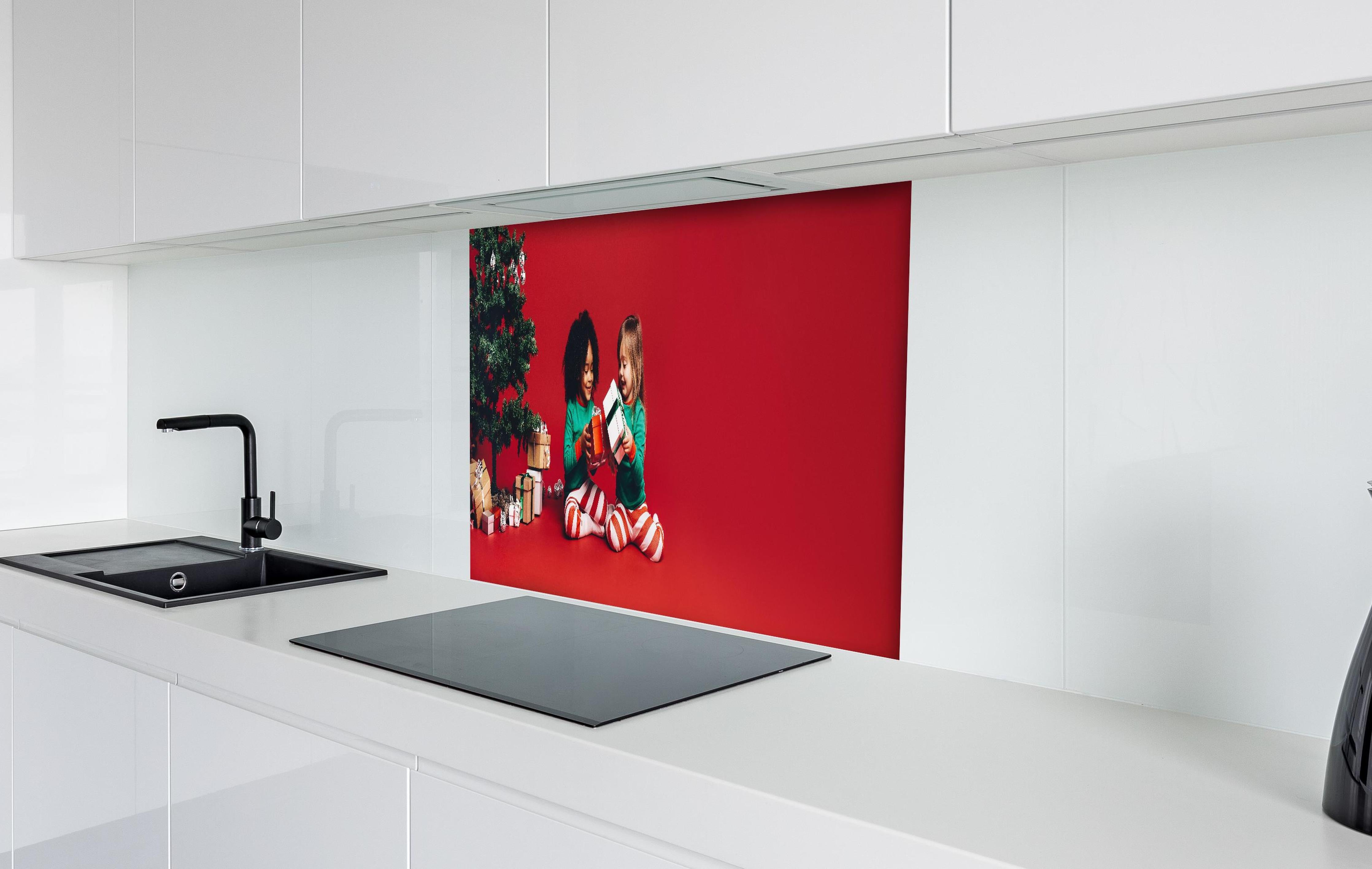 Spritzschutz - Fröhliche Kinder tauschen Weihnachtsgeschenke aus hinter einem Cerankochfeld zwischen Holz-Kochutensilien