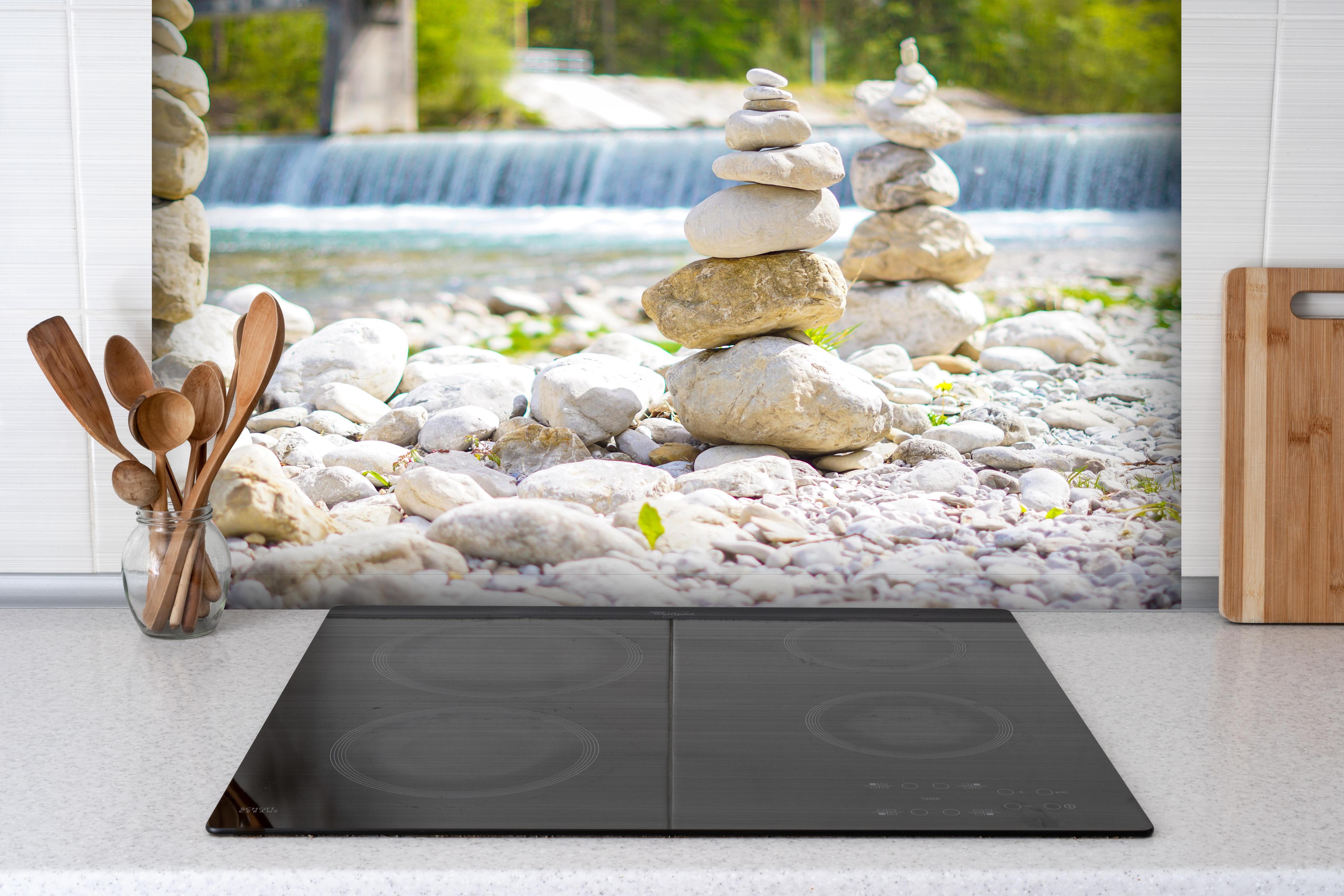 Spritzschutz - Gestapelte Steine am Fluss in Weiß-Grau hinter einem Cerankochfeld zwischen Holz-Kochutensilien