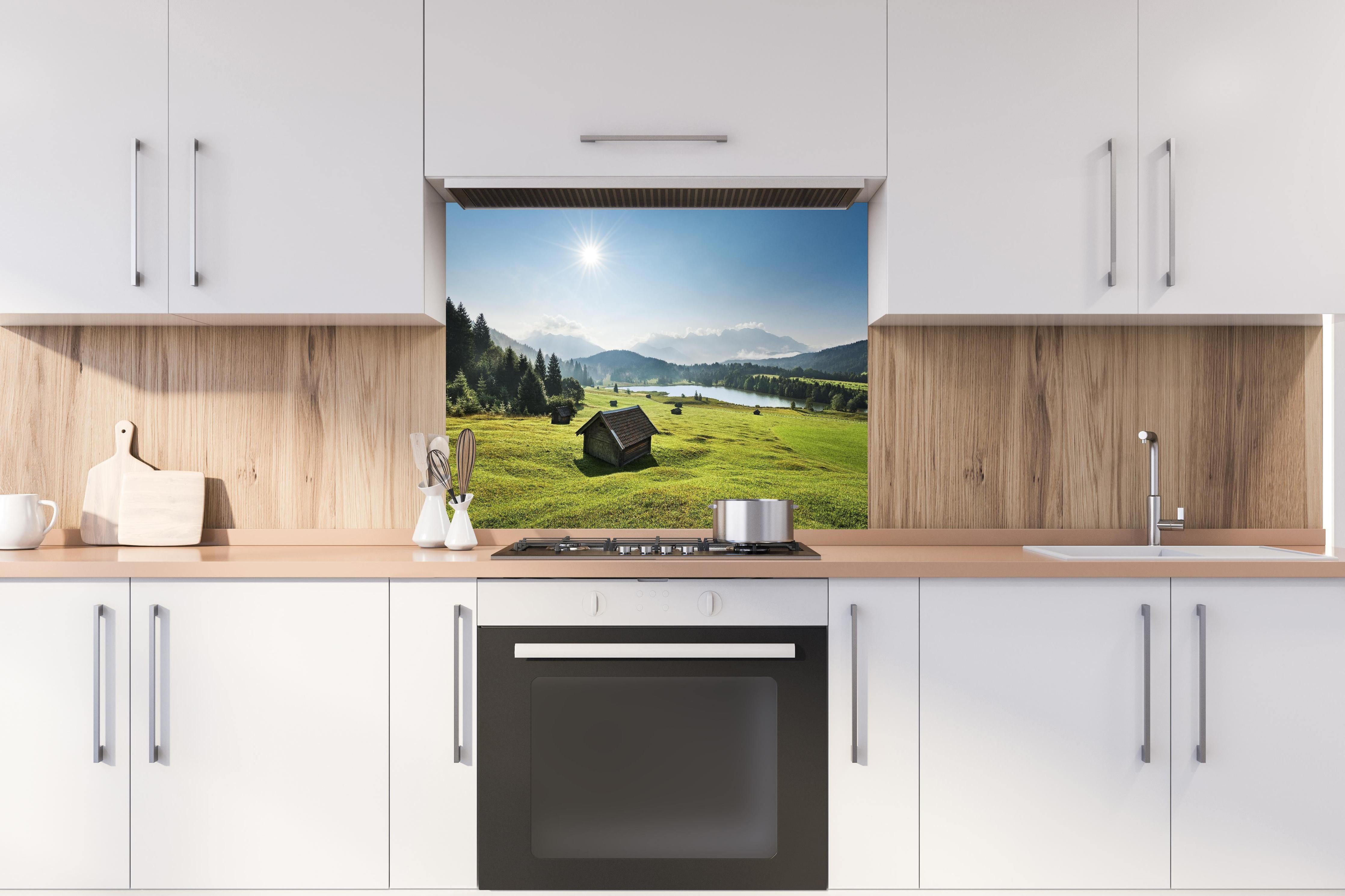 Spritzschutz - Grüne Alpenwiese, Berge und See im Sonnenschein hinter einem Cerankochfeld zwischen Holz-Kochutensilien