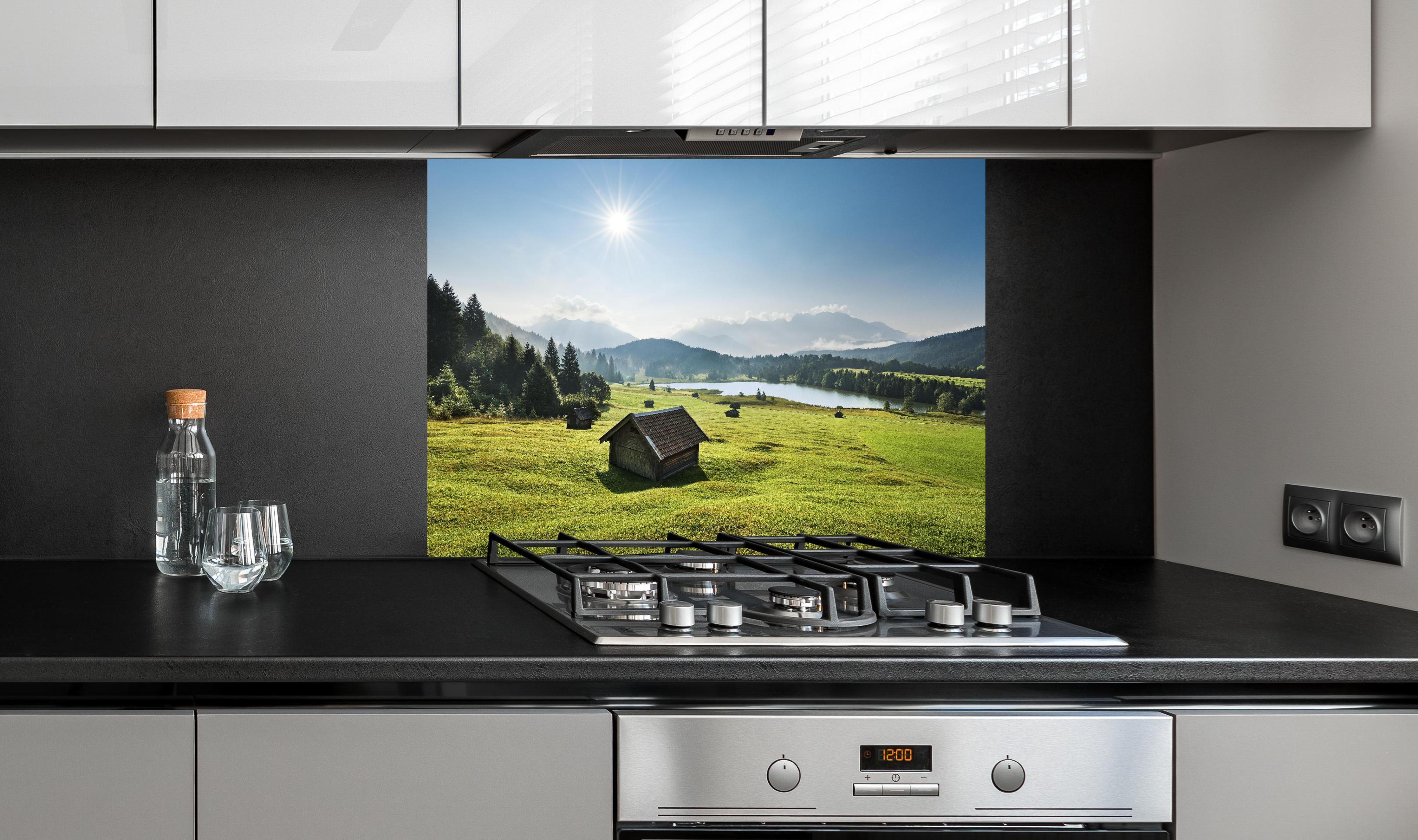 Spritzschutz - Grüne Alpenwiese, Berge und See im Sonnenschein hinter einem Cerankochfeld zwischen Holz-Kochutensilien