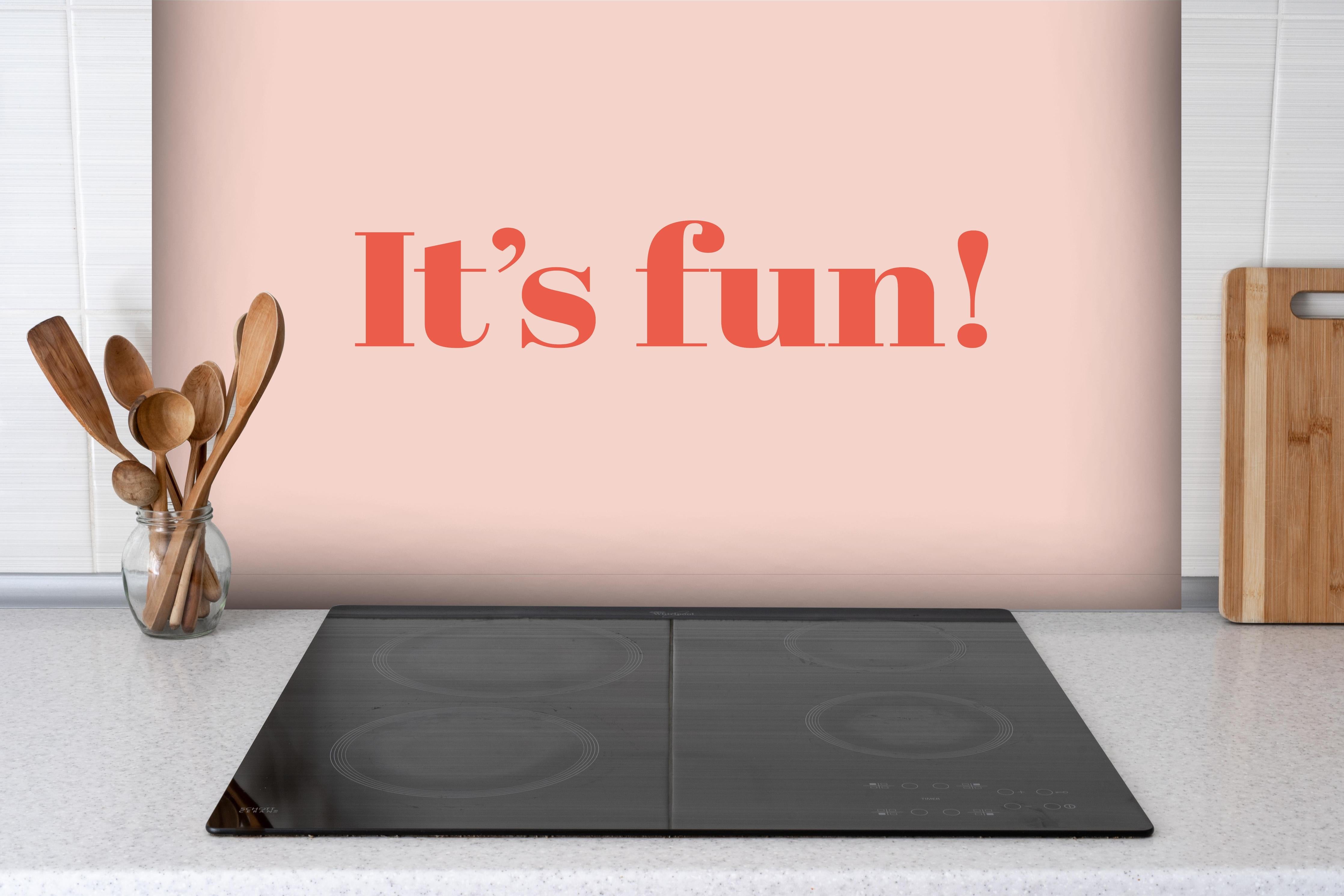 Spritzschutz - Inspirierender 'It's Fun!' Typo-Print in Pink hinter einem Cerankochfeld zwischen Holz-Kochutensilien