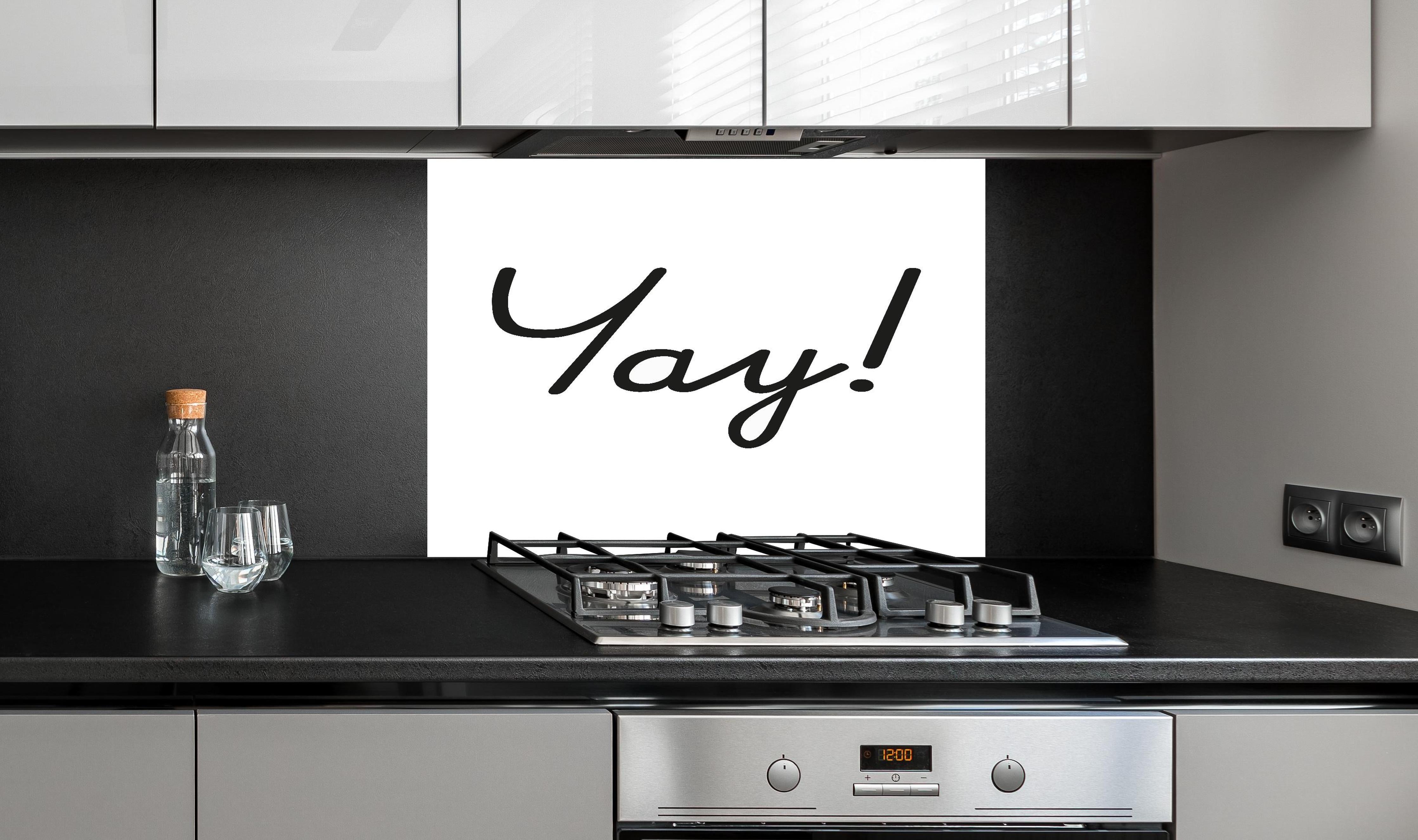 Spritzschutz - Inspirierender 'Yay!' Schriftzug in Schwarz auf Weiß hinter einem Cerankochfeld zwischen Holz-Kochutensilien