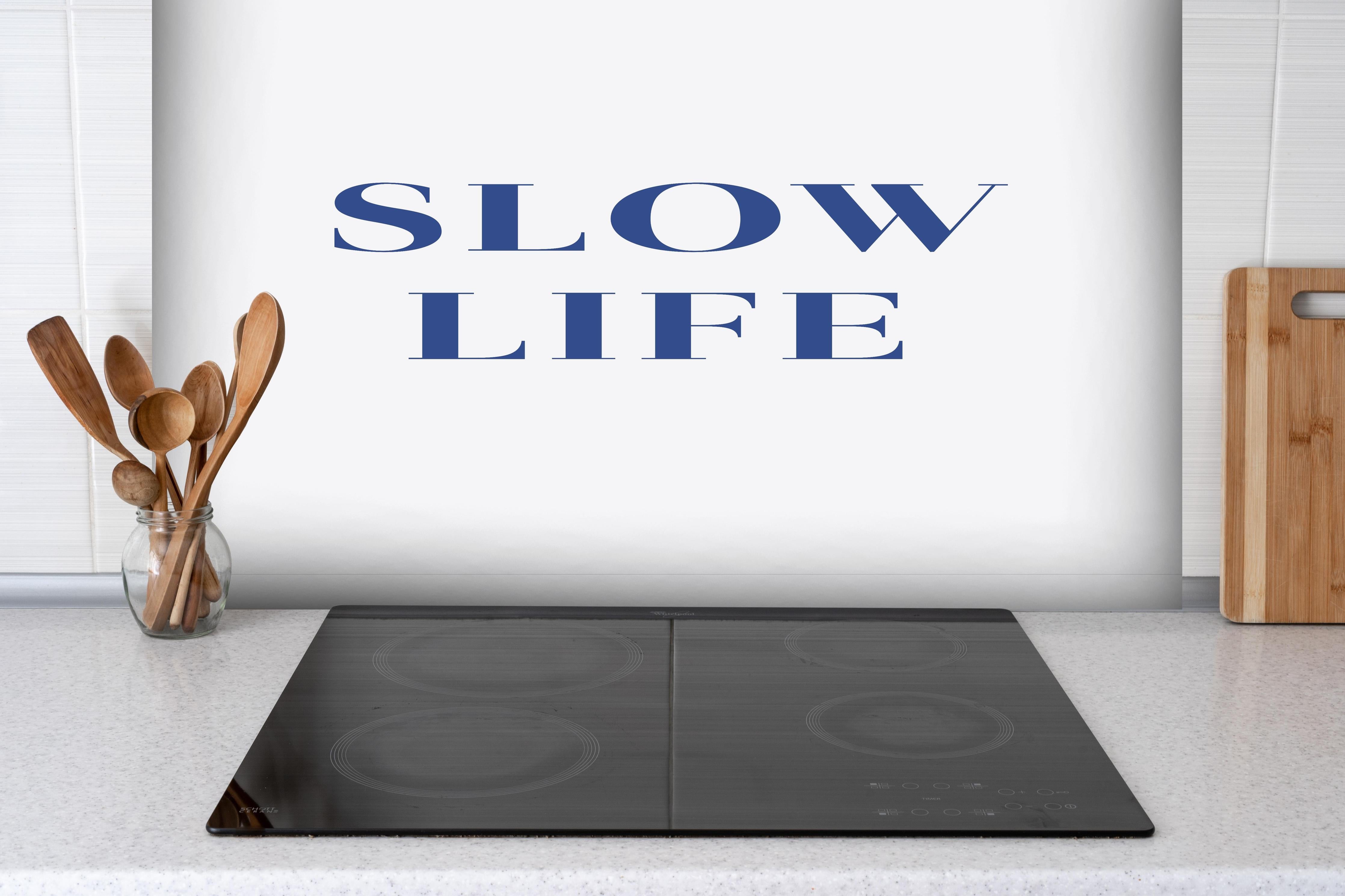 Spritzschutz - Inspirierendes Slow Life Zitat, Blau auf Weiß hinter einem Cerankochfeld zwischen Holz-Kochutensilien