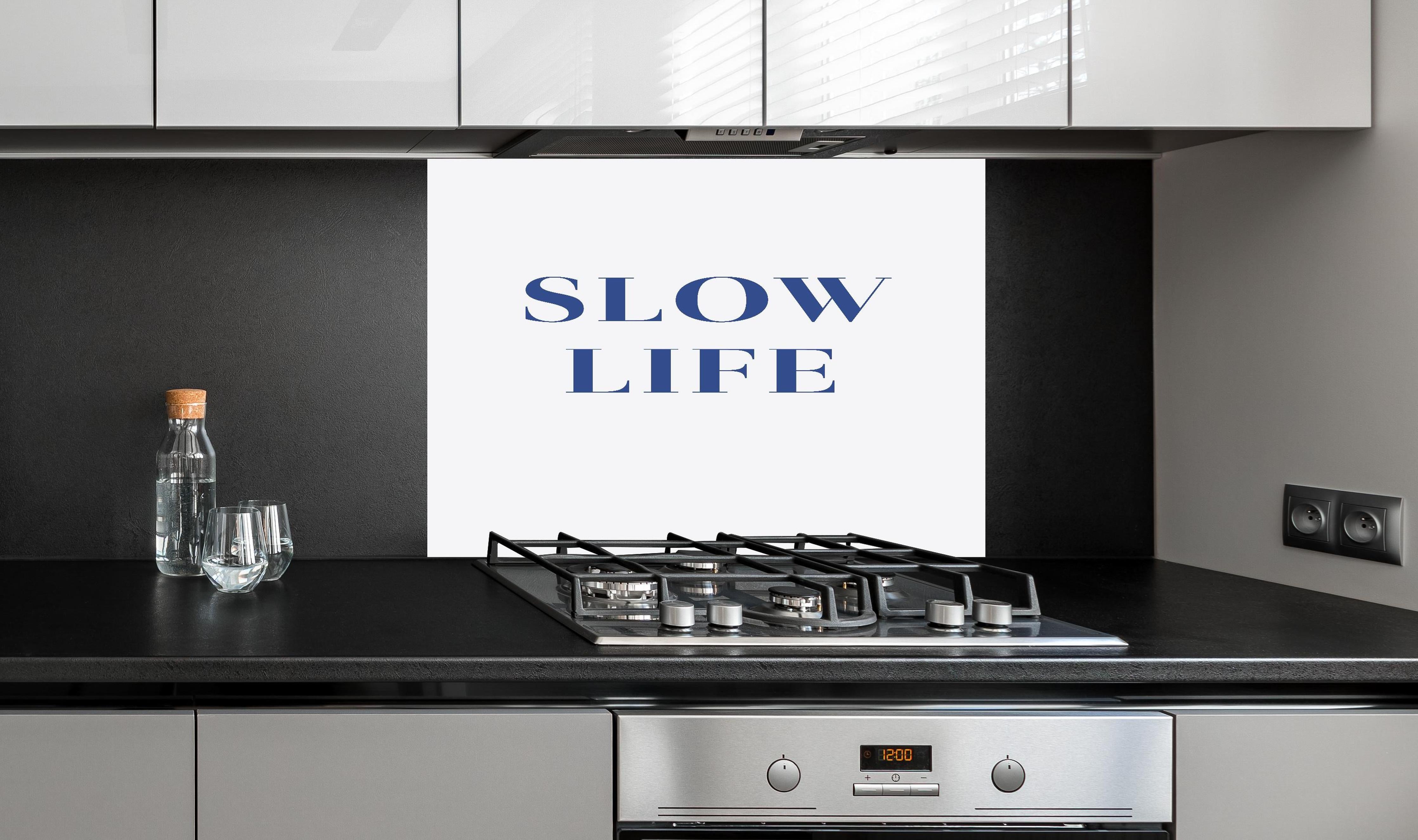 Spritzschutz - Inspirierendes Slow Life Zitat, Blau auf Weiß hinter einem Cerankochfeld zwischen Holz-Kochutensilien
