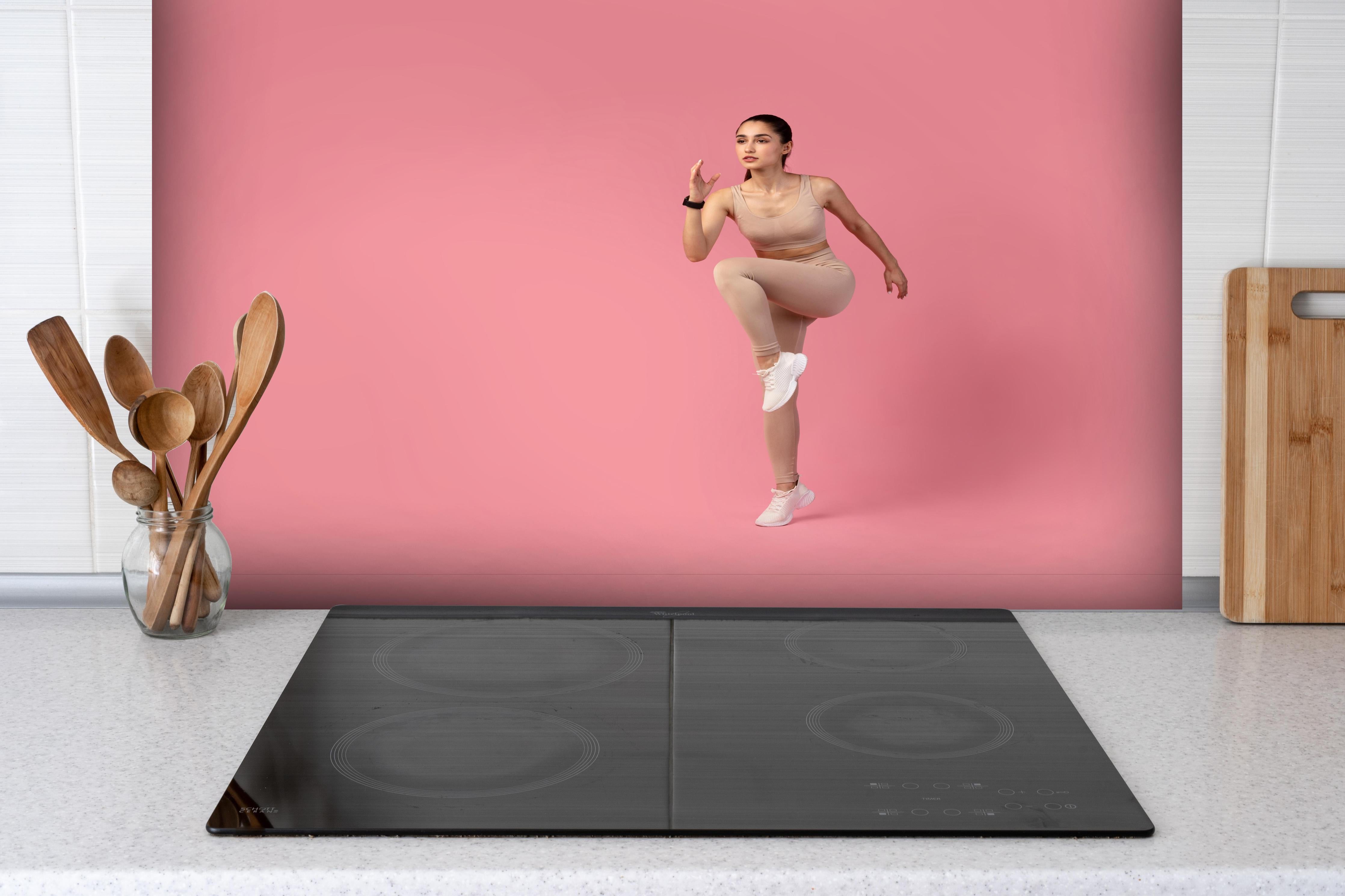 Spritzschutz - Junge Fitness Frau in Läuferpose hinter einem Cerankochfeld zwischen Holz-Kochutensilien