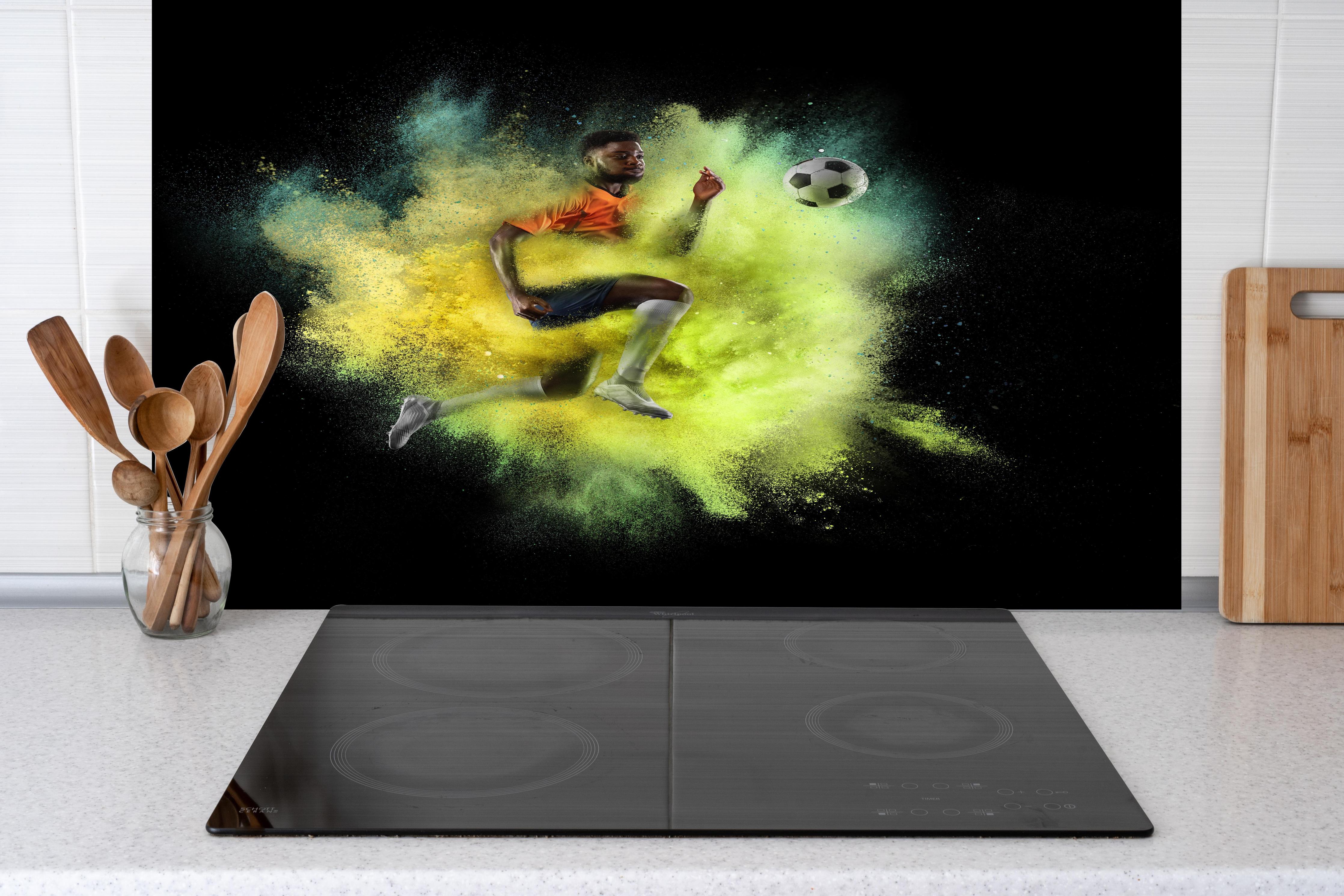 Spritzschutz - Junger Fußballspieler in Farbexplosion hinter einem Cerankochfeld zwischen Holz-Kochutensilien