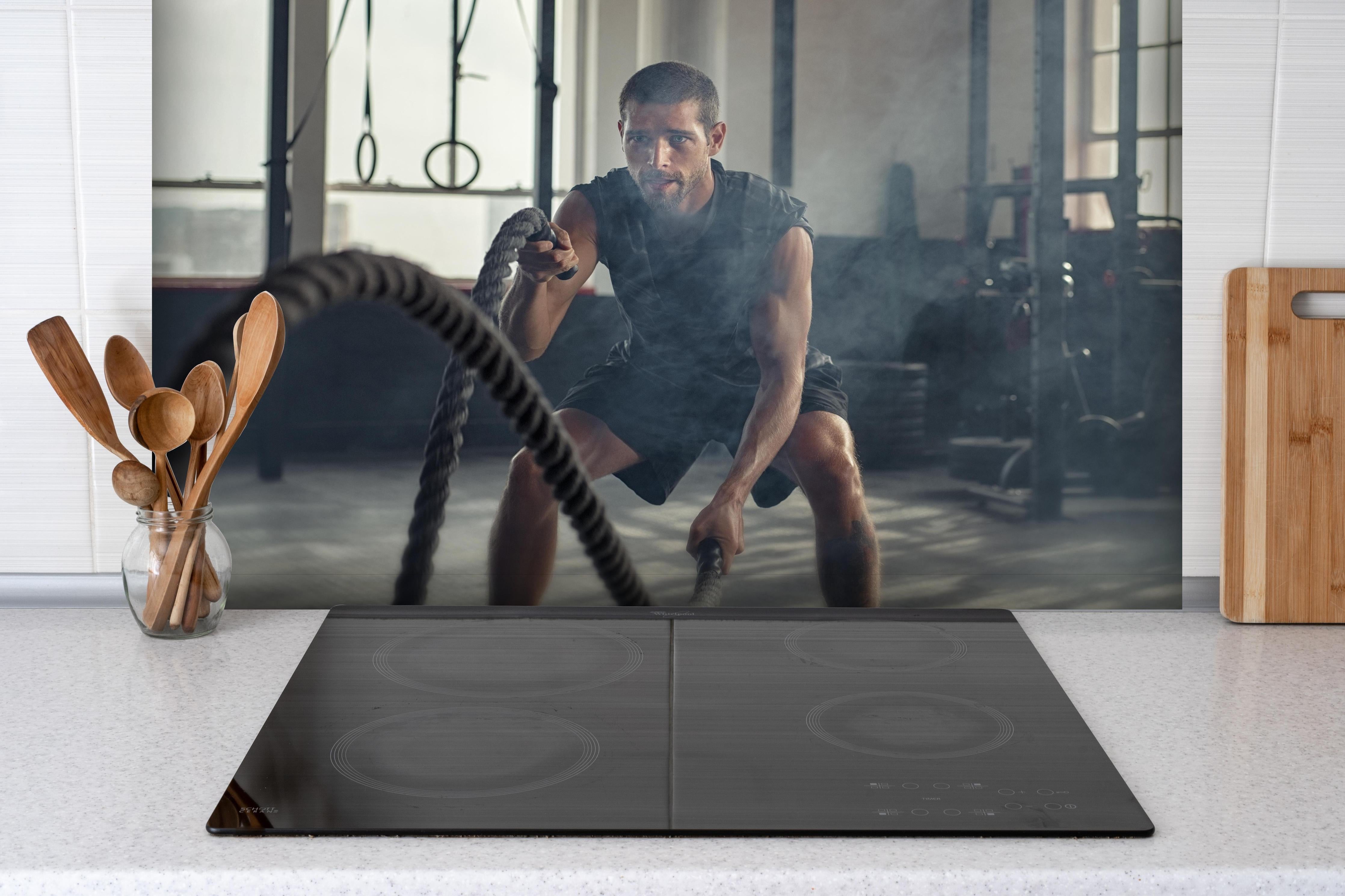 Spritzschutz - Junger Mann der trainiert hinter einem Cerankochfeld zwischen Holz-Kochutensilien