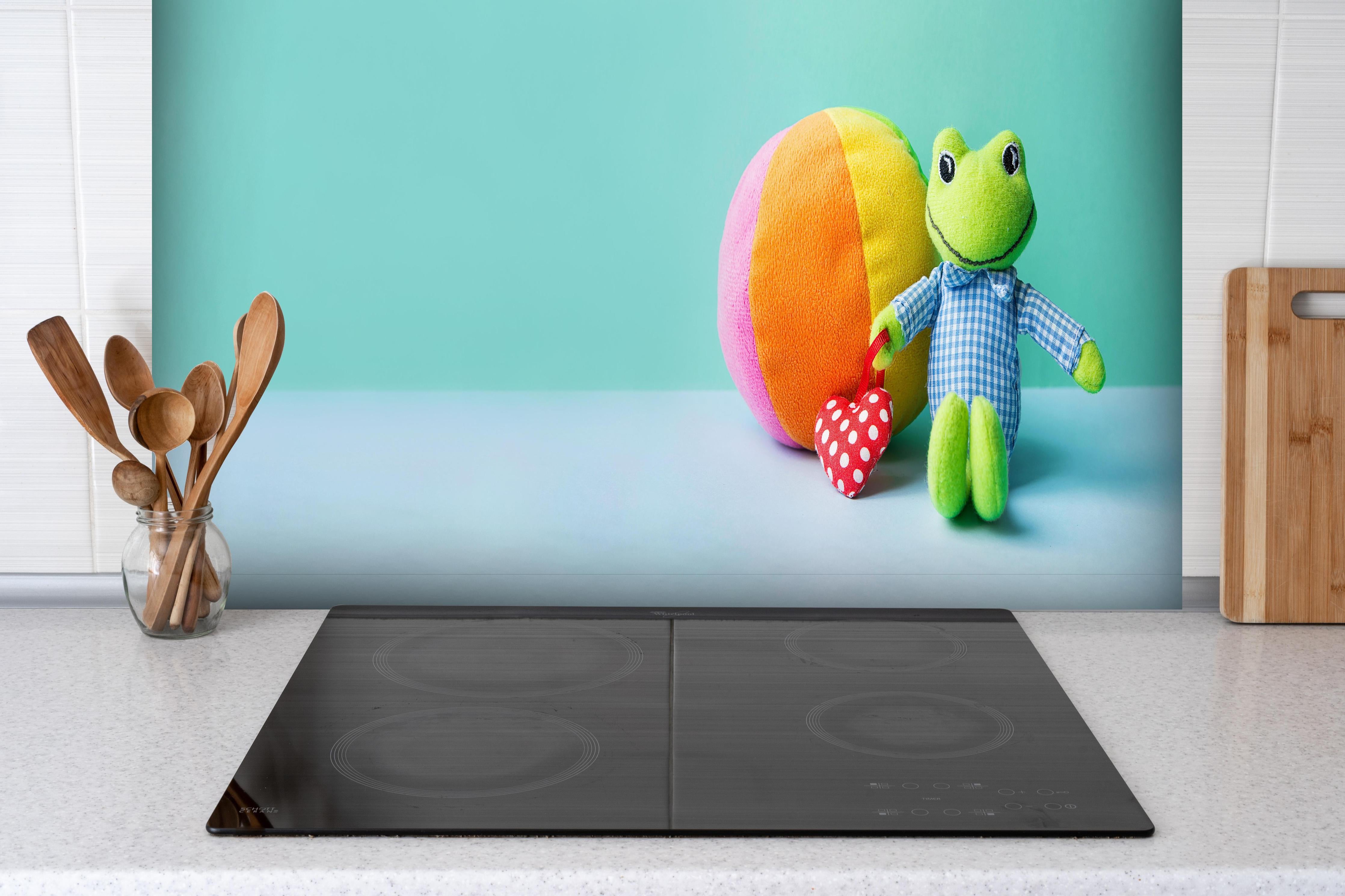 Spritzschutz - Kinderspielzeug Kleiner Frosch hinter einem Cerankochfeld zwischen Holz-Kochutensilien