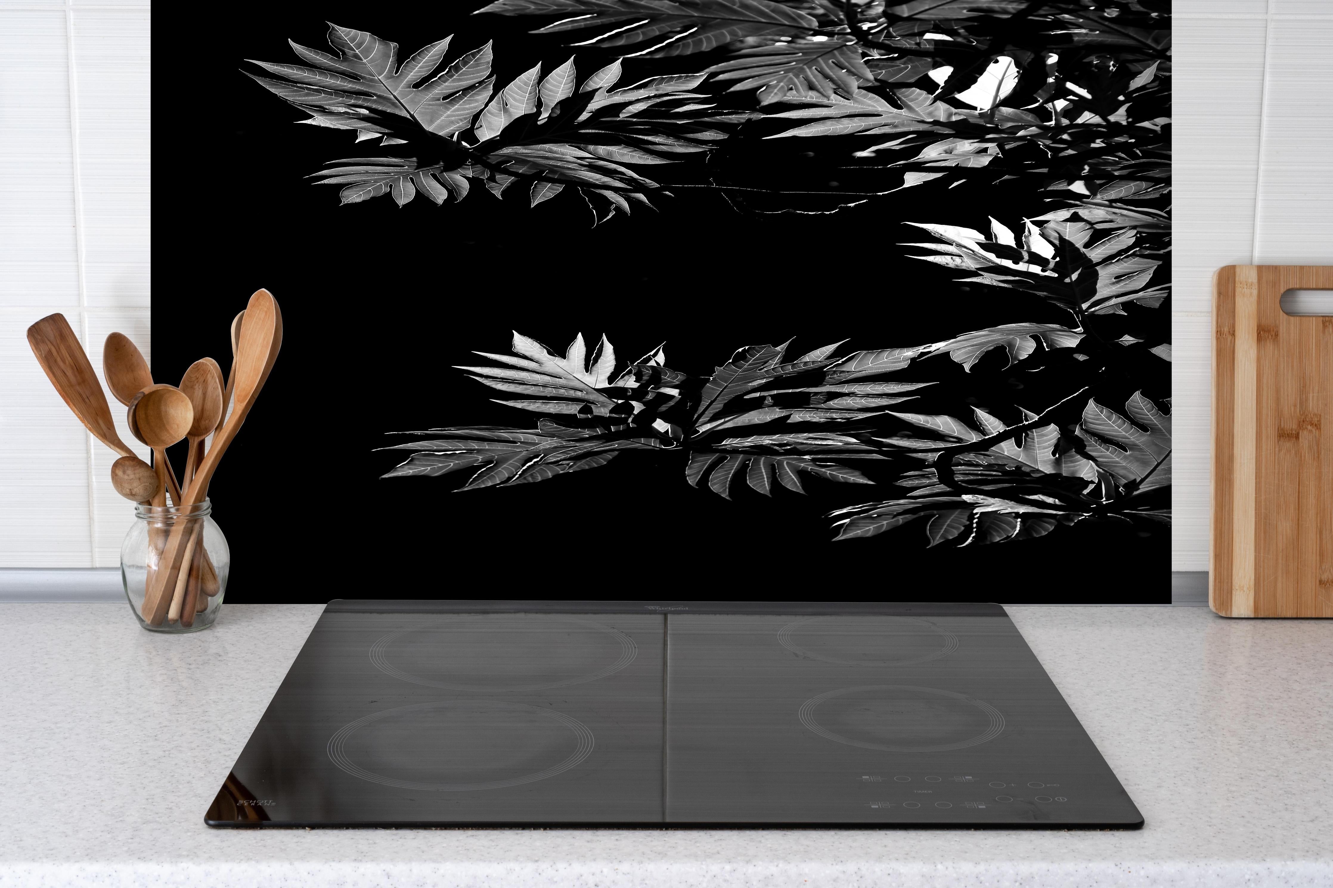 Spritzschutz - Kontrastreiche Blätter auf schwarzem Hintergrund hinter einem Cerankochfeld zwischen Holz-Kochutensilien