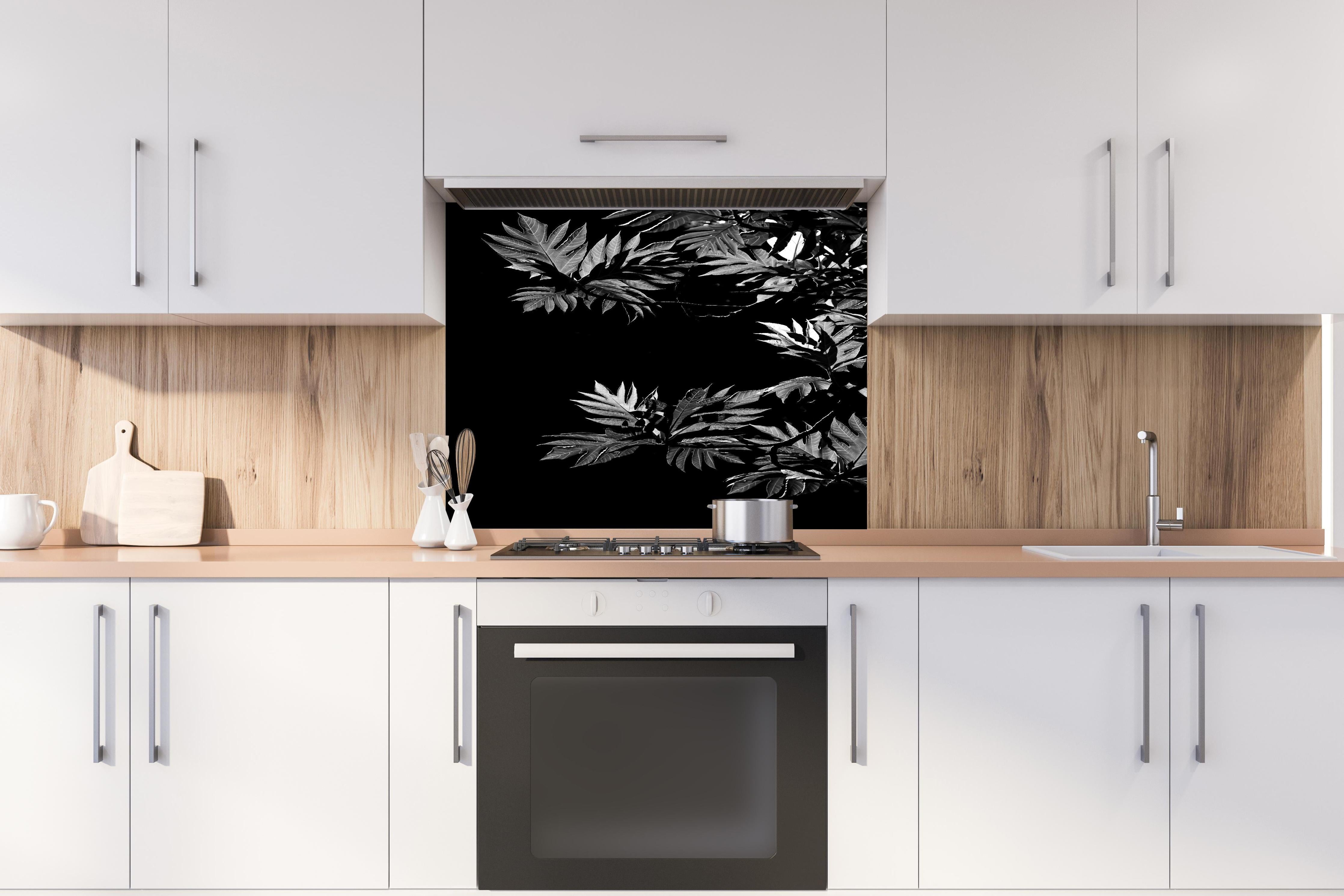 Spritzschutz - Kontrastreiche Blätter auf schwarzem Hintergrund hinter einem Cerankochfeld zwischen Holz-Kochutensilien