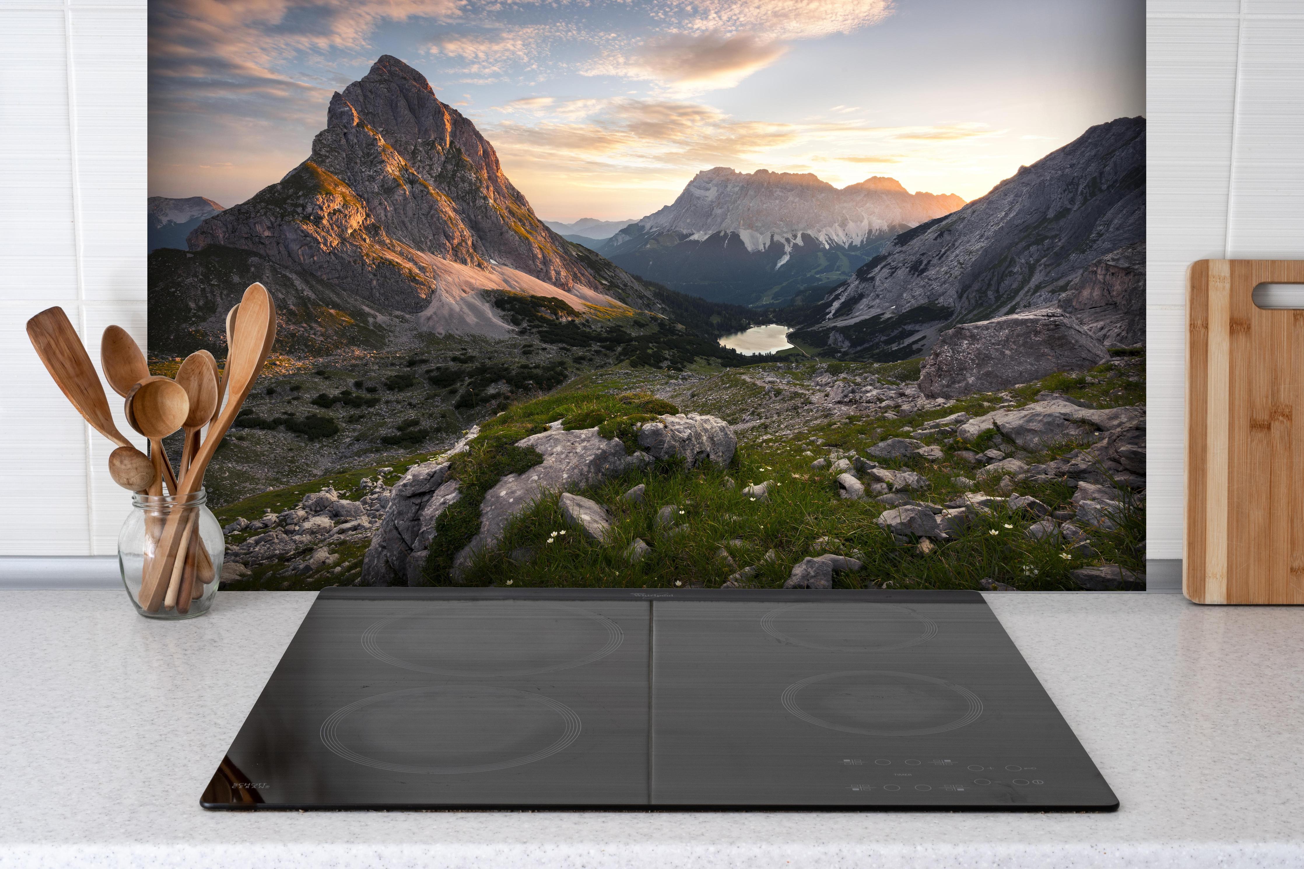 Spritzschutz - Malerische Alpenlandschaft bei Sonnenuntergang hinter einem Cerankochfeld zwischen Holz-Kochutensilien