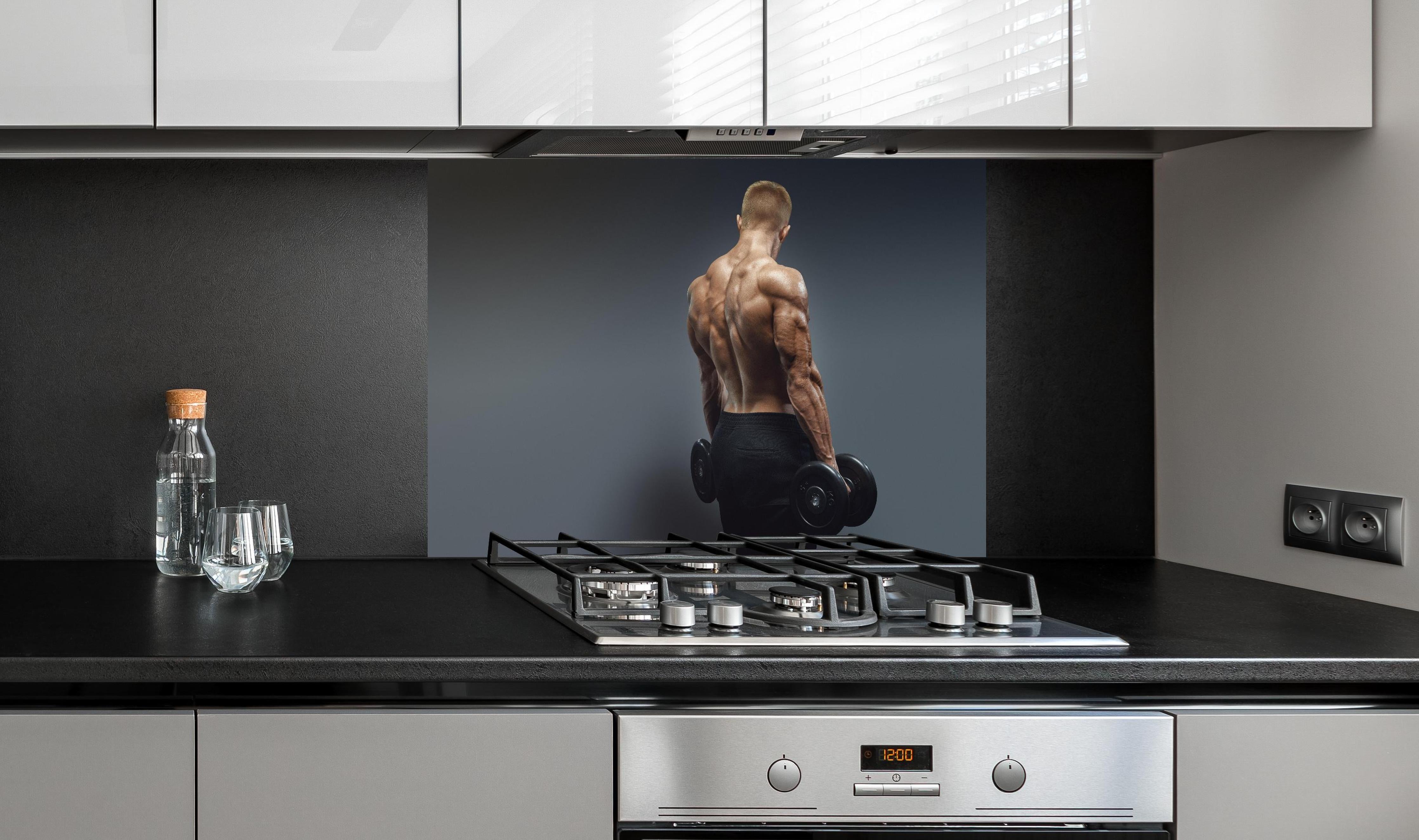 Spritzschutz - Muskulöser Sportler mit Hanteln hinter einem Cerankochfeld zwischen Holz-Kochutensilien