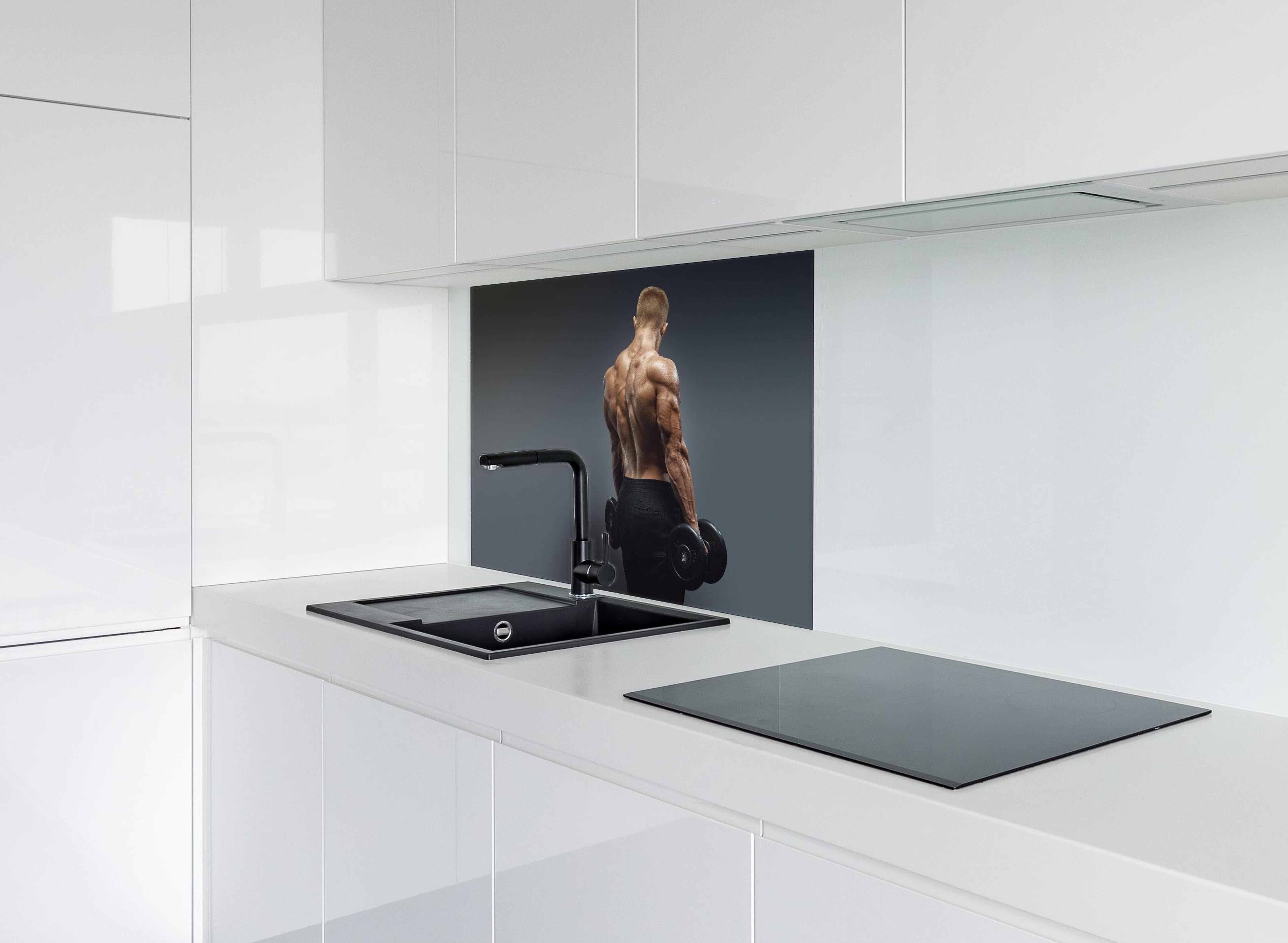 Spritzschutz - Muskulöser Sportler mit Hanteln hinter einem Cerankochfeld zwischen Holz-Kochutensilien