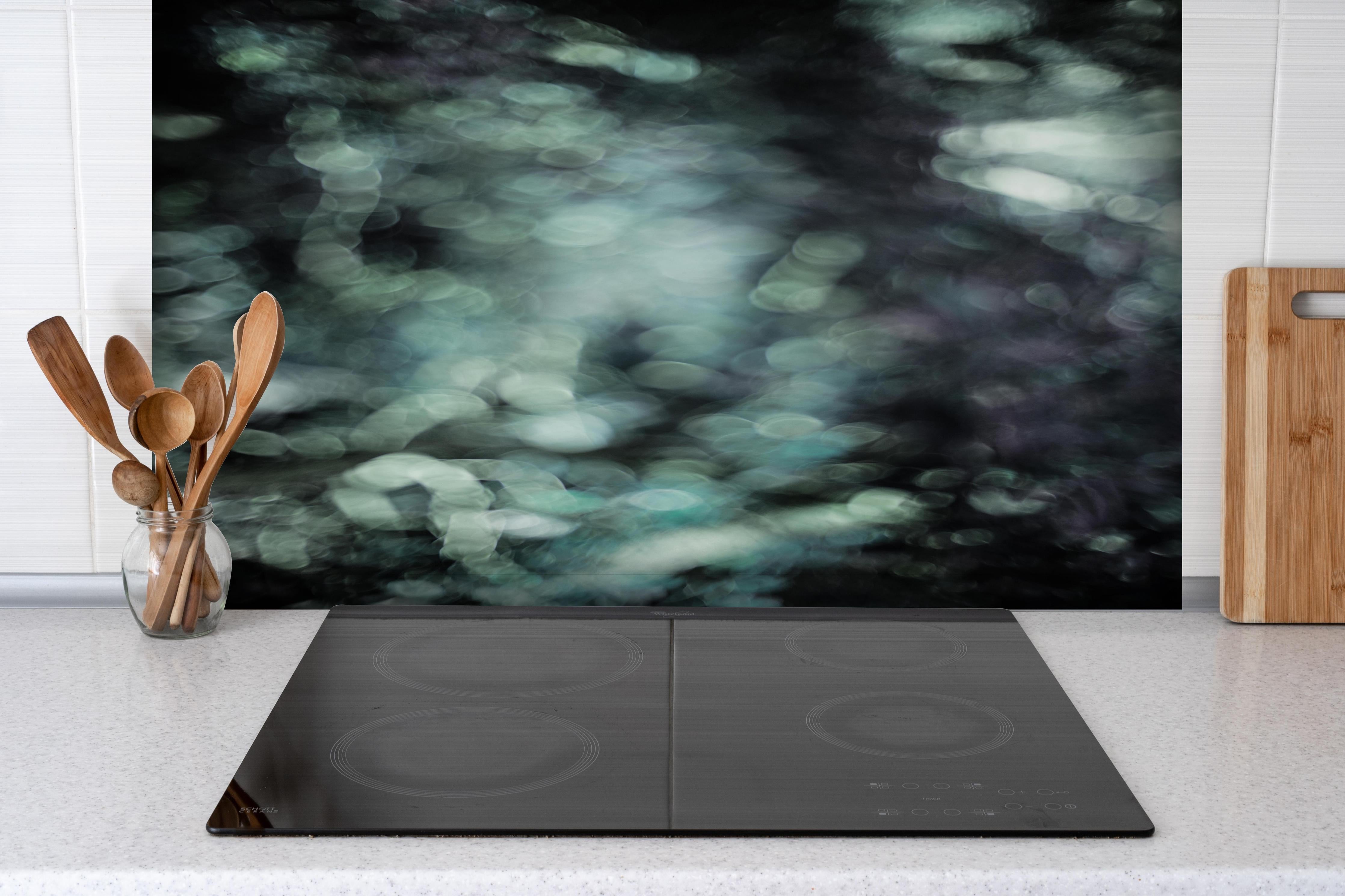 Spritzschutz - Mystischer Grünschimmer auf dunklem Grund hinter einem Cerankochfeld zwischen Holz-Kochutensilien