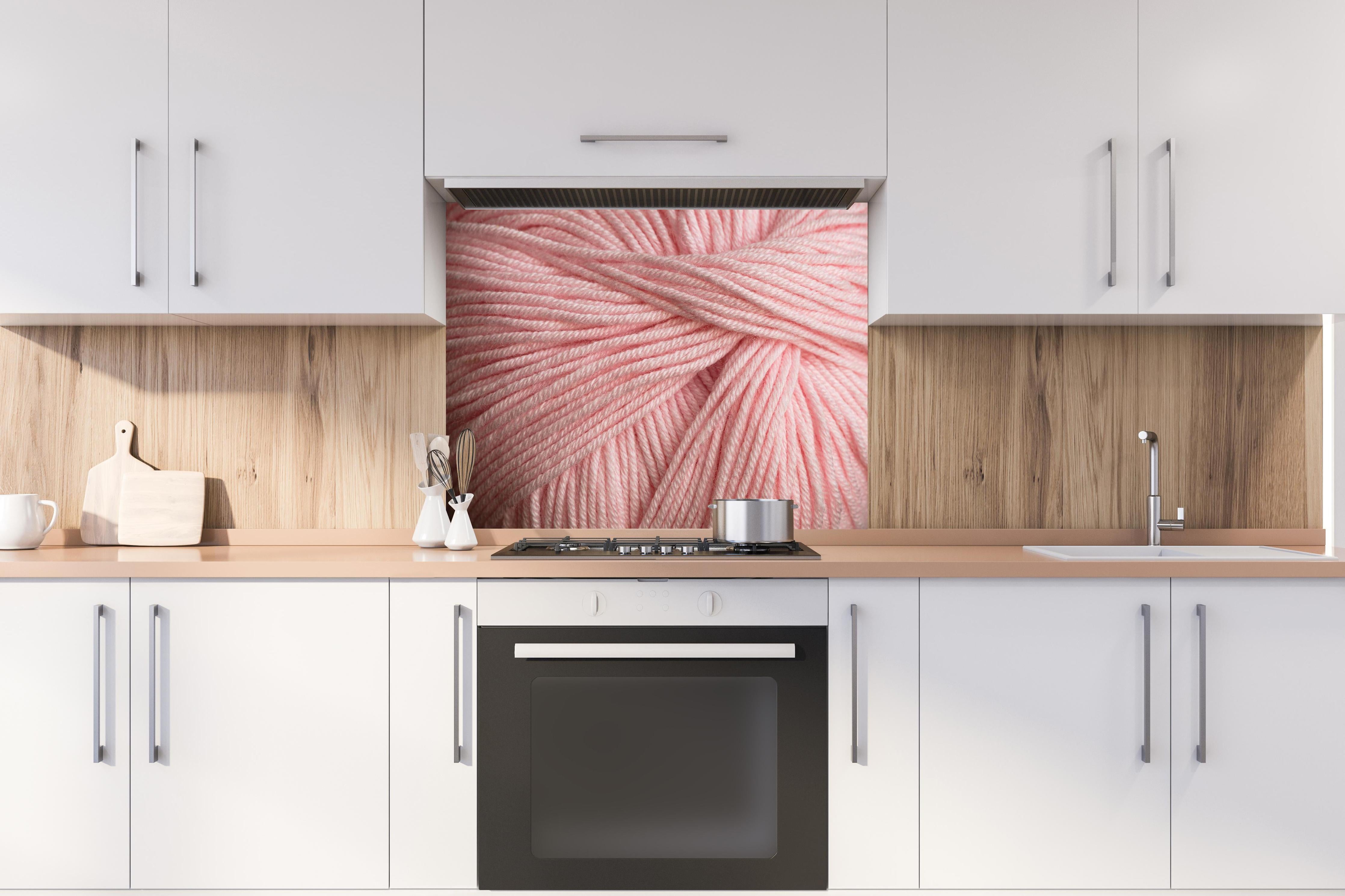 Spritzschutz - Nahaufnahme eines rosa Garnknäuels hinter einem Cerankochfeld zwischen Holz-Kochutensilien