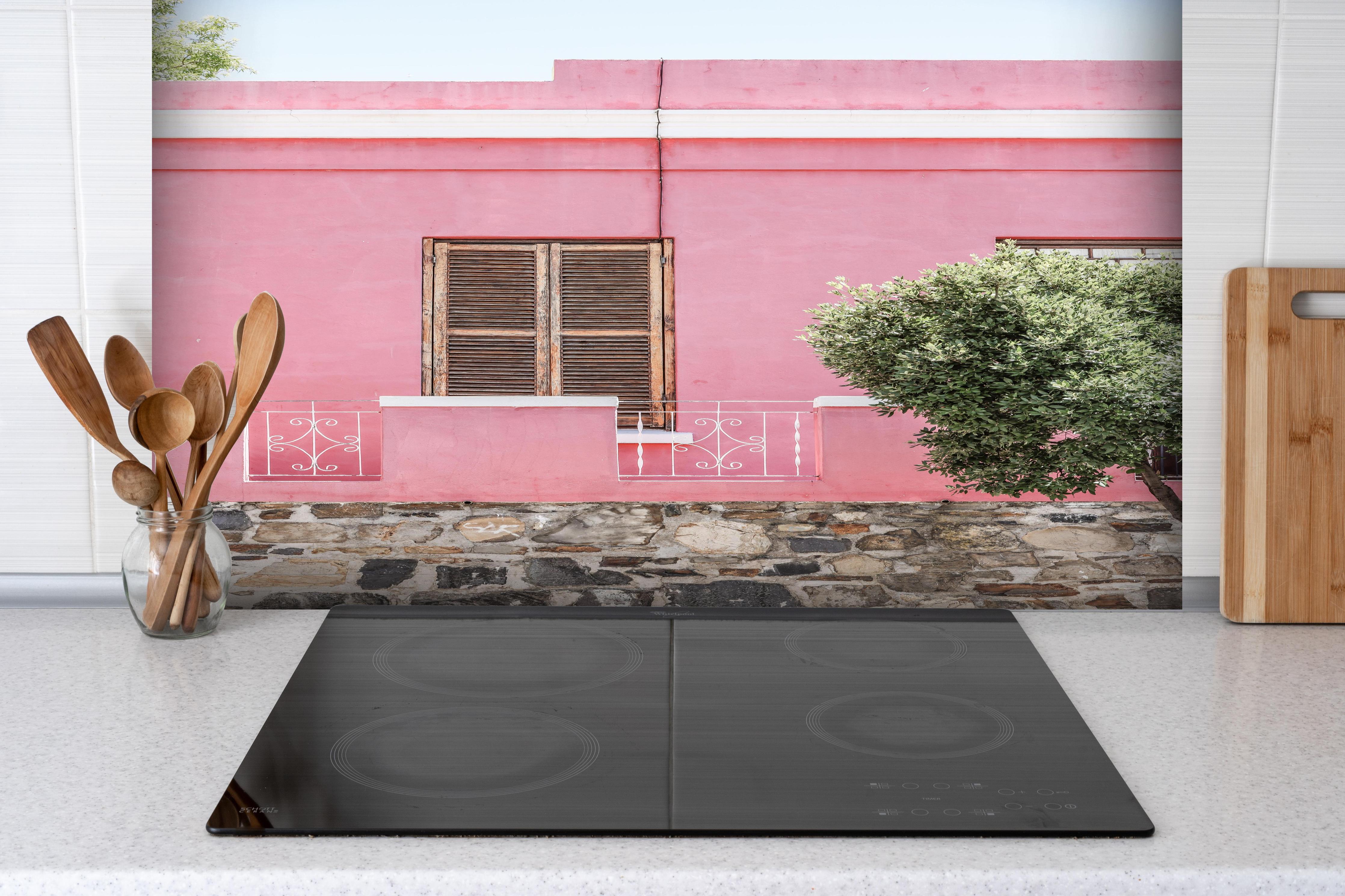 Spritzschutz - Pinkes Haus mit Baum und Steinwand hinter einem Cerankochfeld zwischen Holz-Kochutensilien