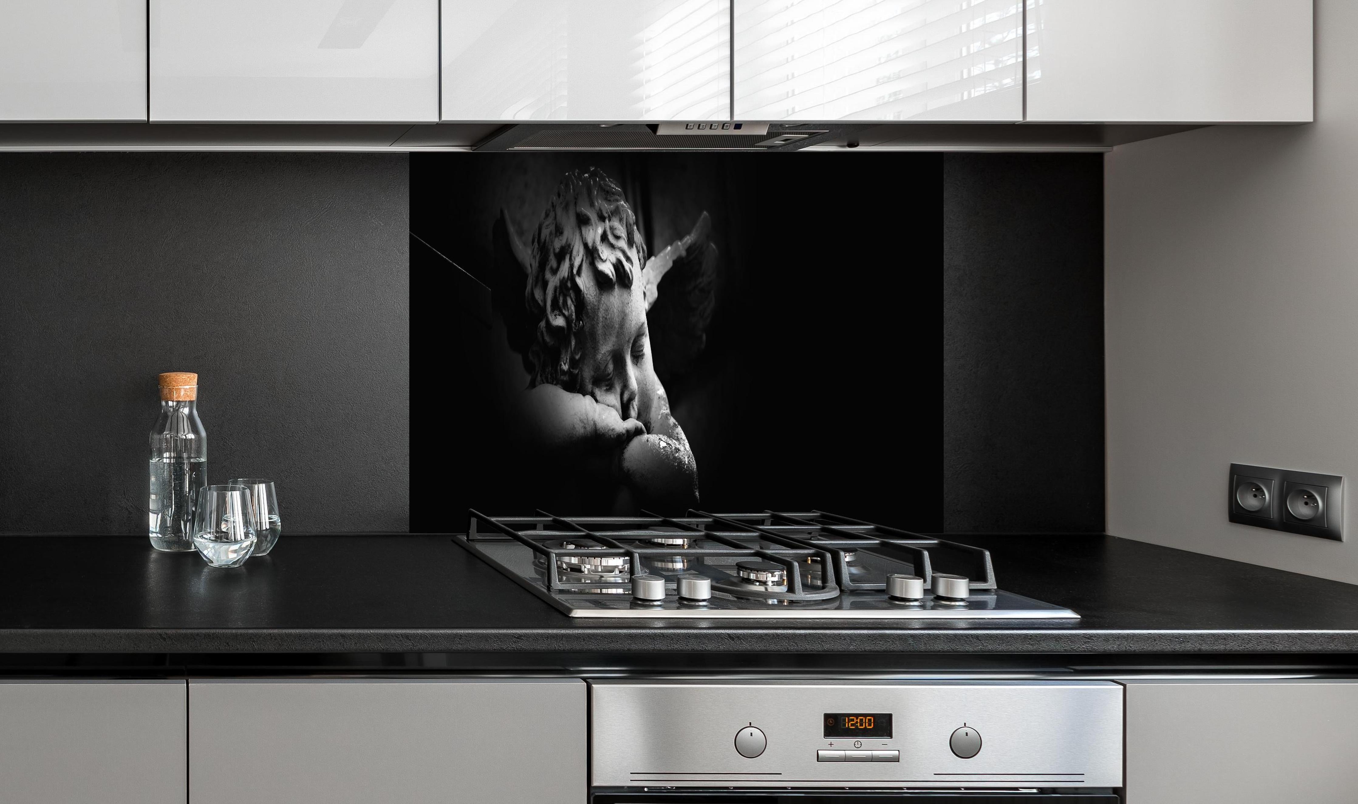 Spritzschutz - Schlafender Engel Statue in Schwarz-Weiß hinter einem Cerankochfeld zwischen Holz-Kochutensilien