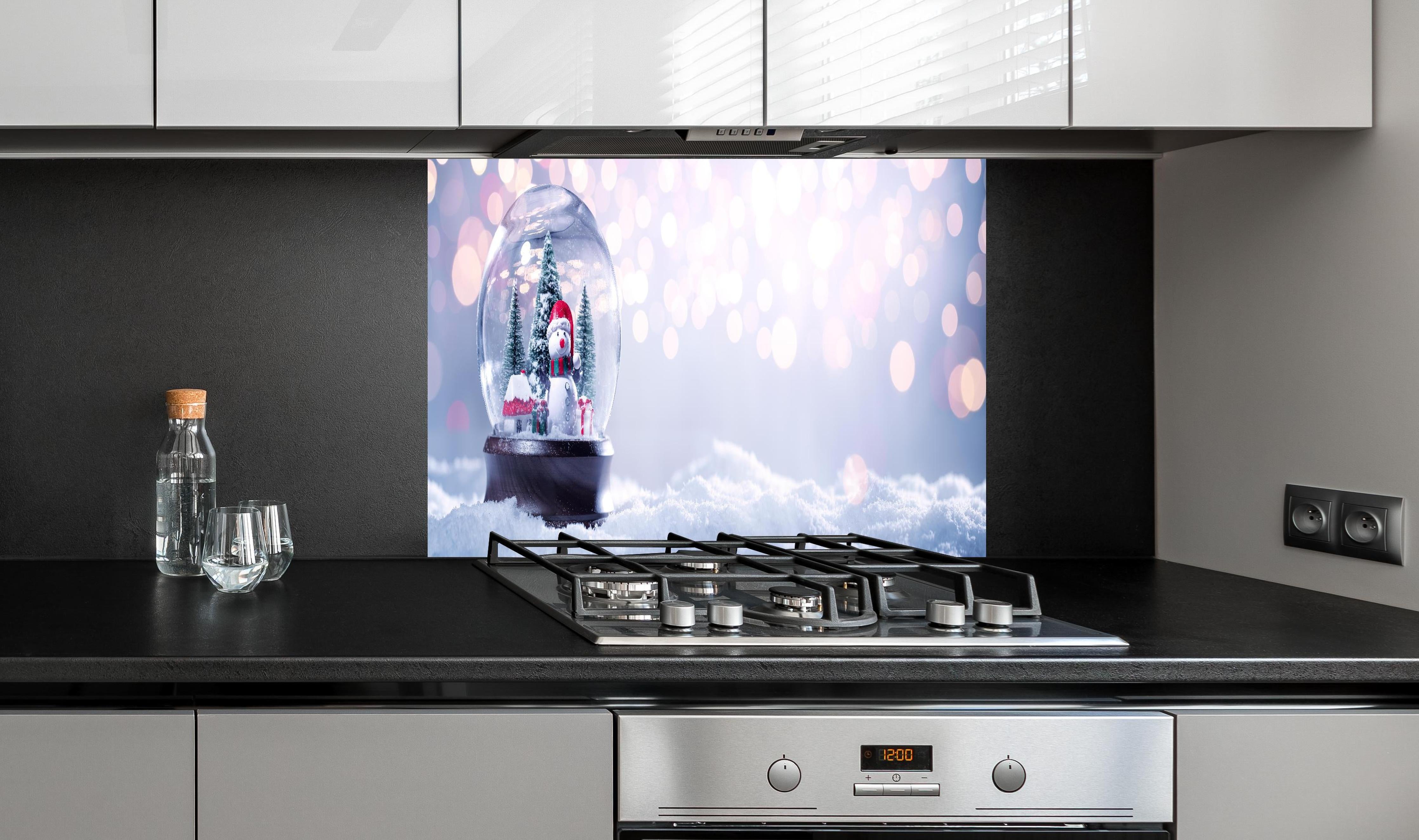 Spritzschutz - Schneekugel auf festlichem Hintergrund hinter einem Cerankochfeld zwischen Holz-Kochutensilien