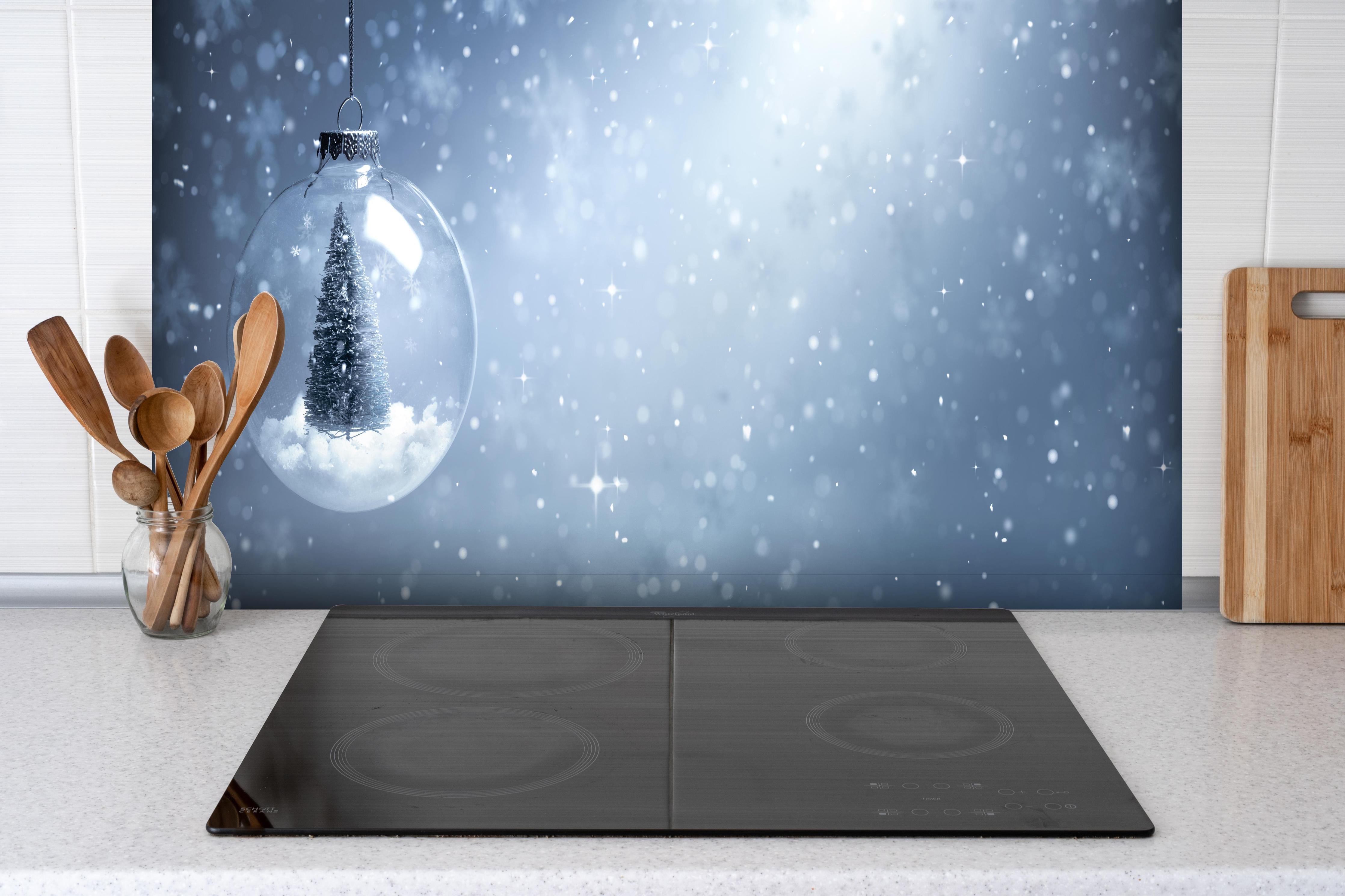 Spritzschutz - Schneekugel mit Weihnachtsbaum hinter einem Cerankochfeld zwischen Holz-Kochutensilien