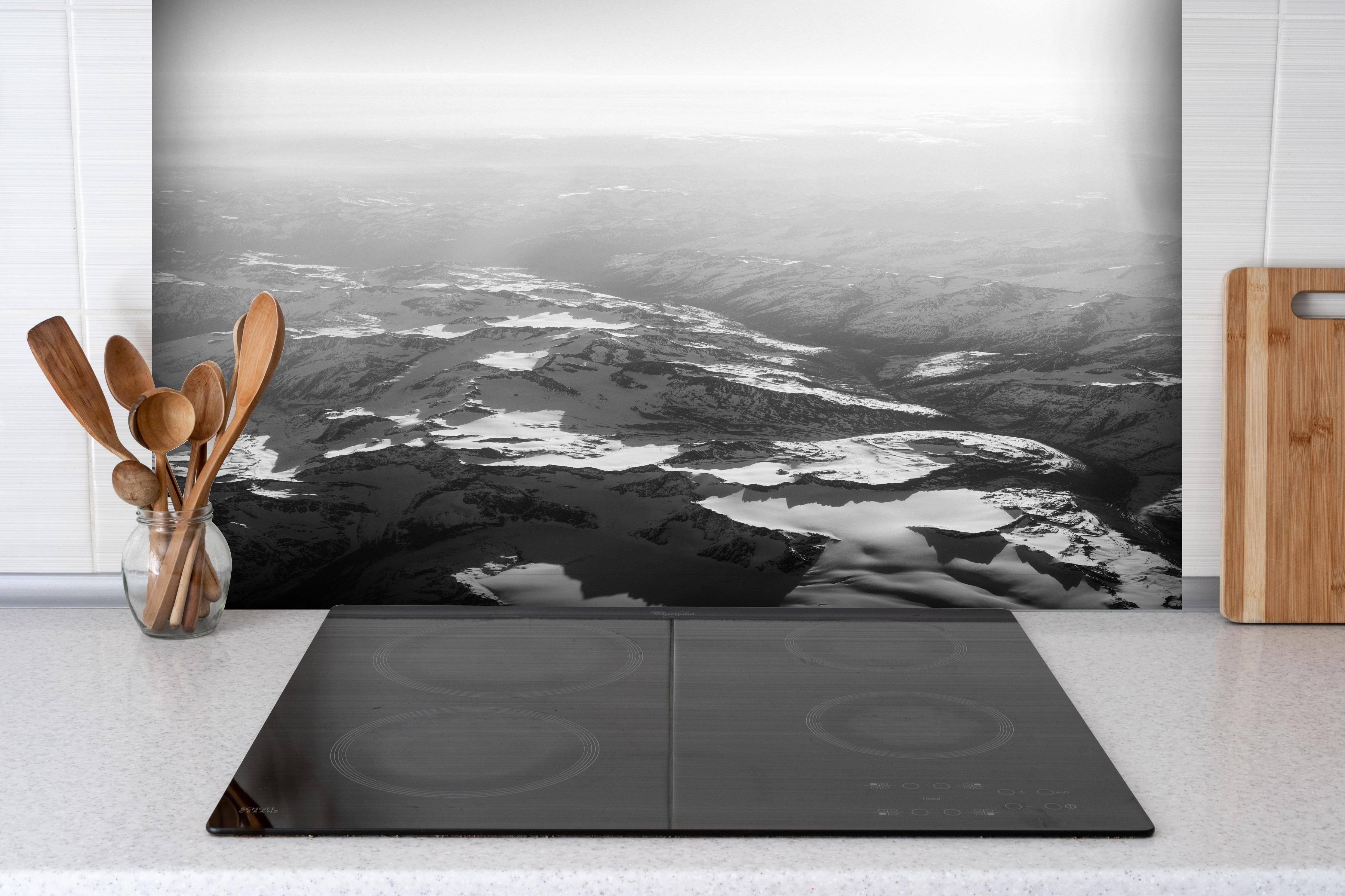 Spritzschutz - Schwarz-Weiß Fotografie Schneebedeckte Alpen hinter einem Cerankochfeld zwischen Holz-Kochutensilien