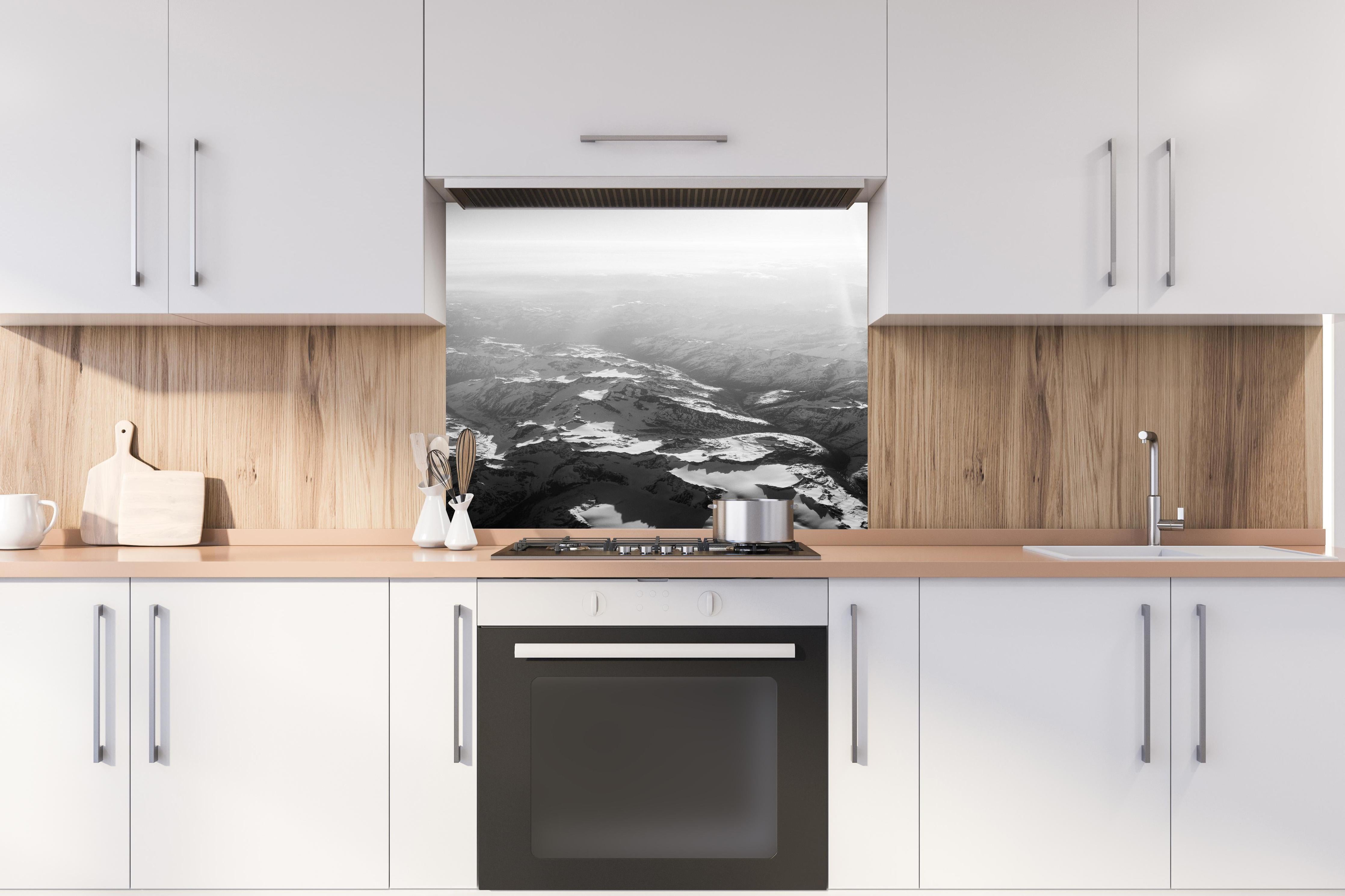 Spritzschutz - Schwarz-Weiß Fotografie Schneebedeckte Alpen hinter einem Cerankochfeld zwischen Holz-Kochutensilien