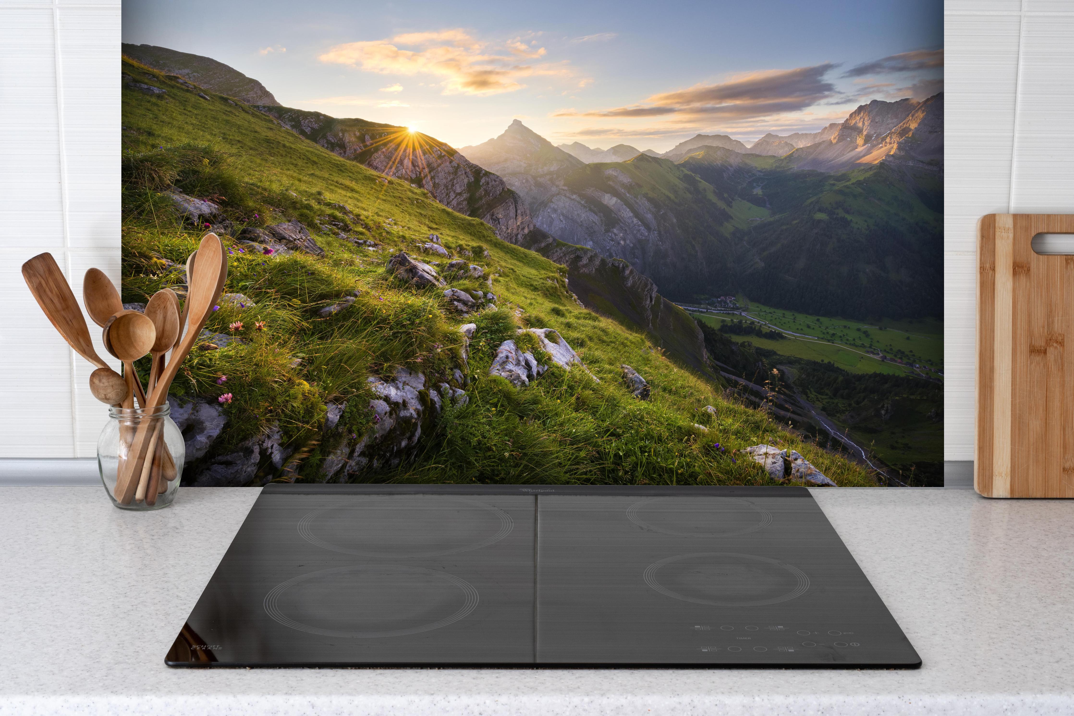 Spritzschutz - Sonnenstrahlen über blühender Alpenwiese hinter einem Cerankochfeld zwischen Holz-Kochutensilien