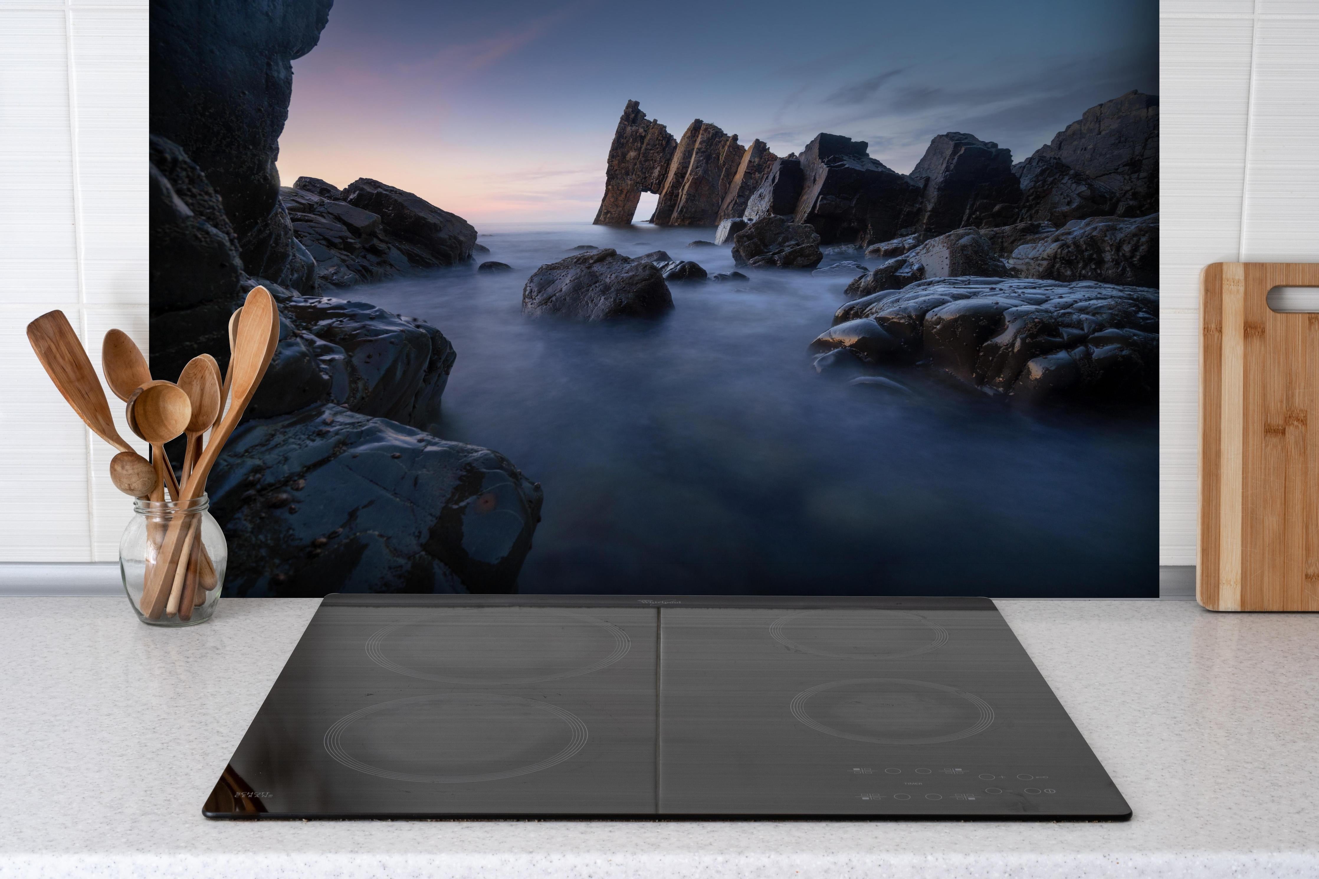 Spritzschutz - Sonnenuntergang am felsigen Meeresufer hinter einem Cerankochfeld zwischen Holz-Kochutensilien