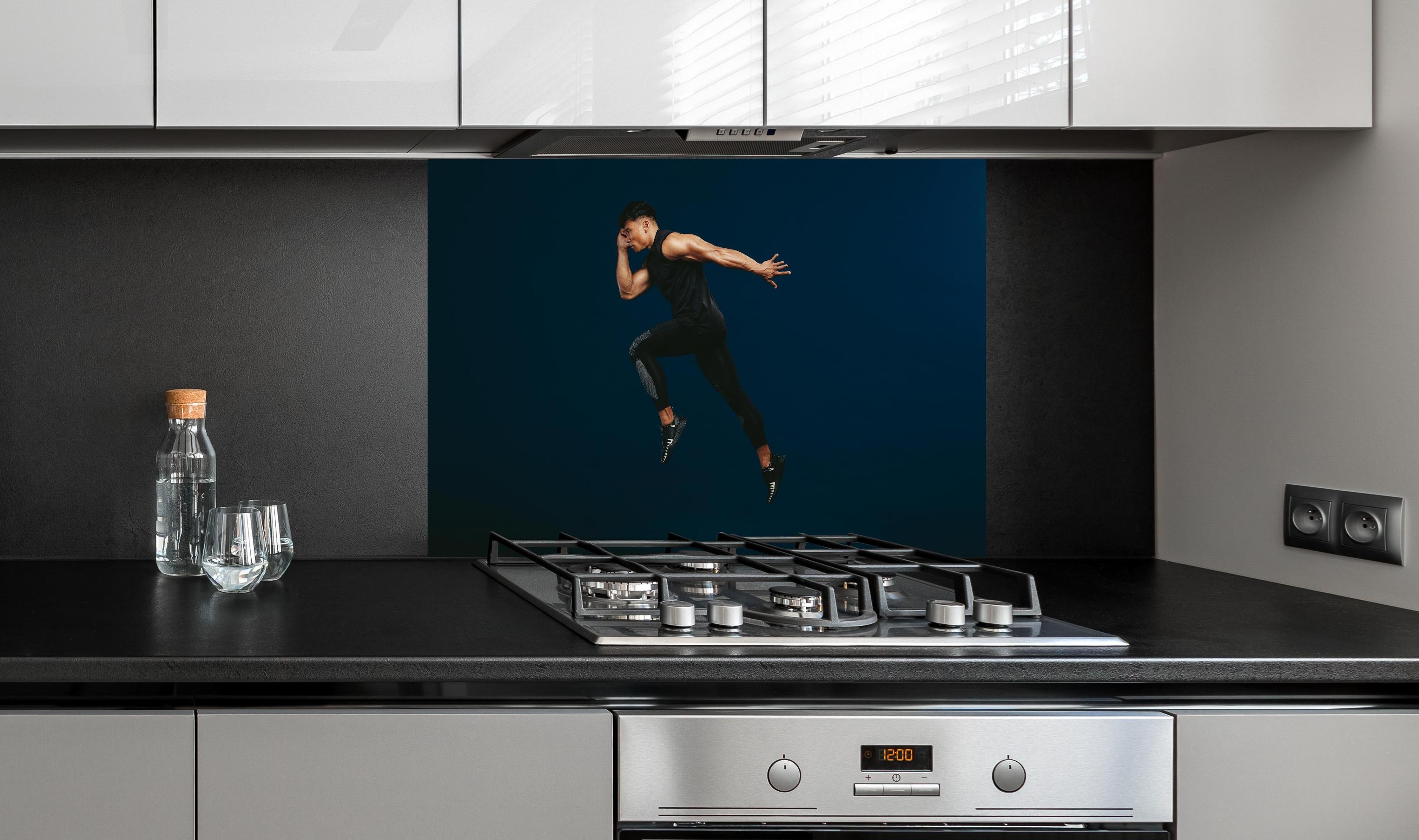 Spritzschutz - Sportler bei einem Sprung hinter einem Cerankochfeld zwischen Holz-Kochutensilien