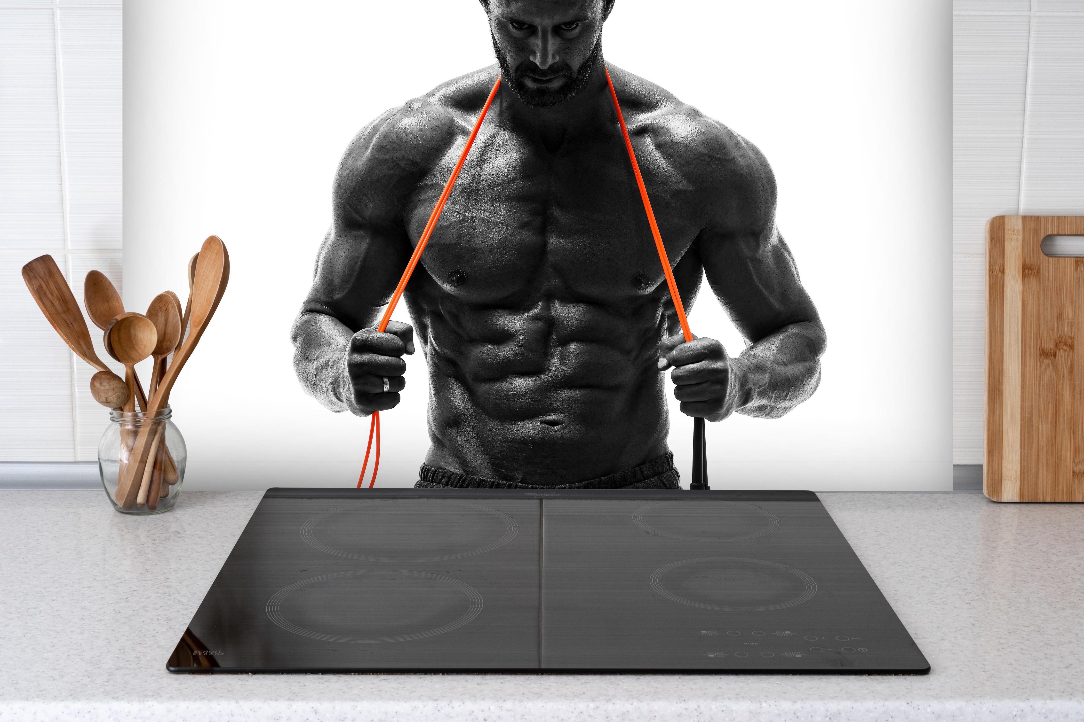 Spritzschutz - Athletischer Mann posiert mit Sprungseil hinter einem Cerankochfeld zwischen Holz-Kochutensilien