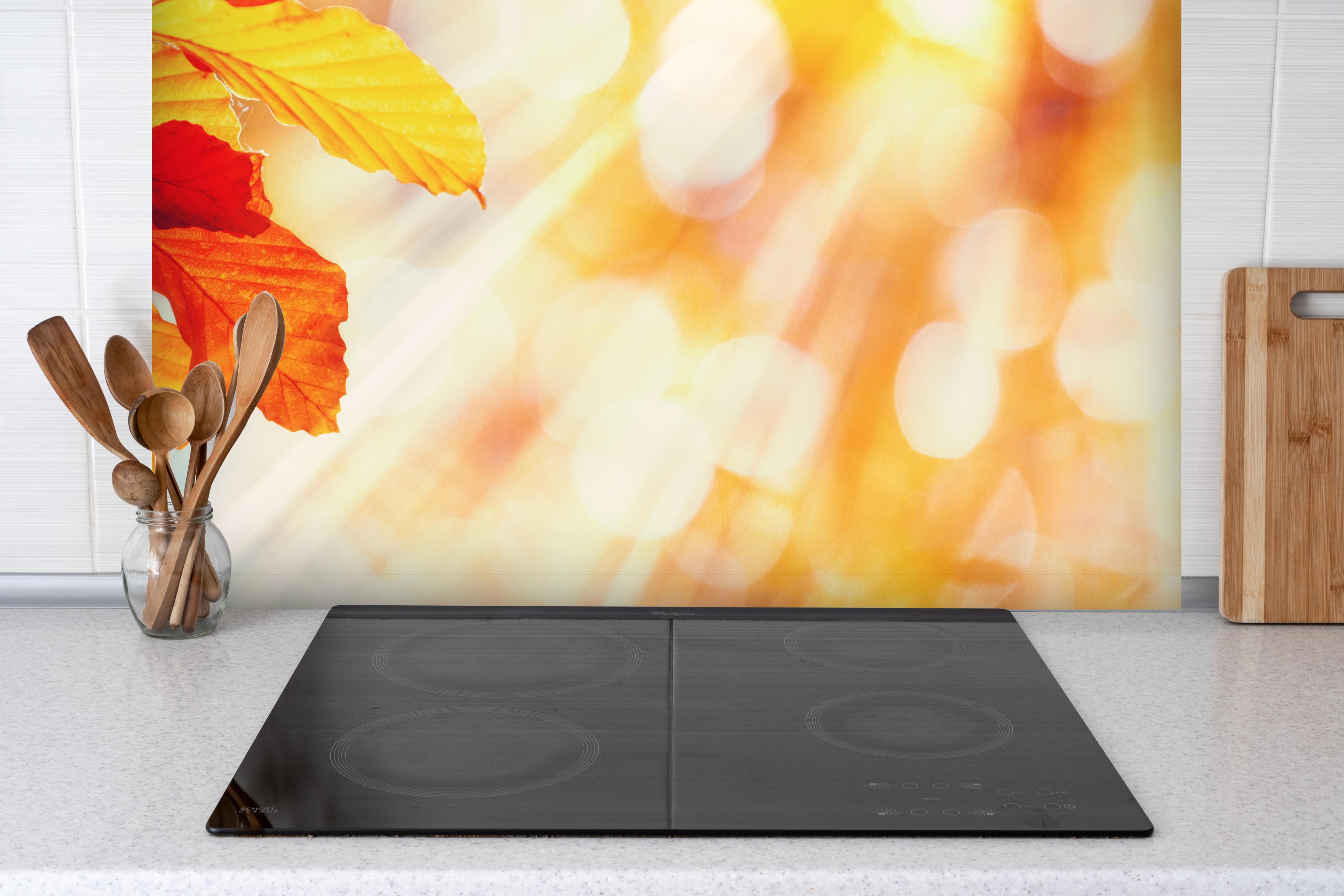 Spritzschutz - Buchenblätter im Sonnenschein hinter einem Cerankochfeld zwischen Holz-Kochutensilien