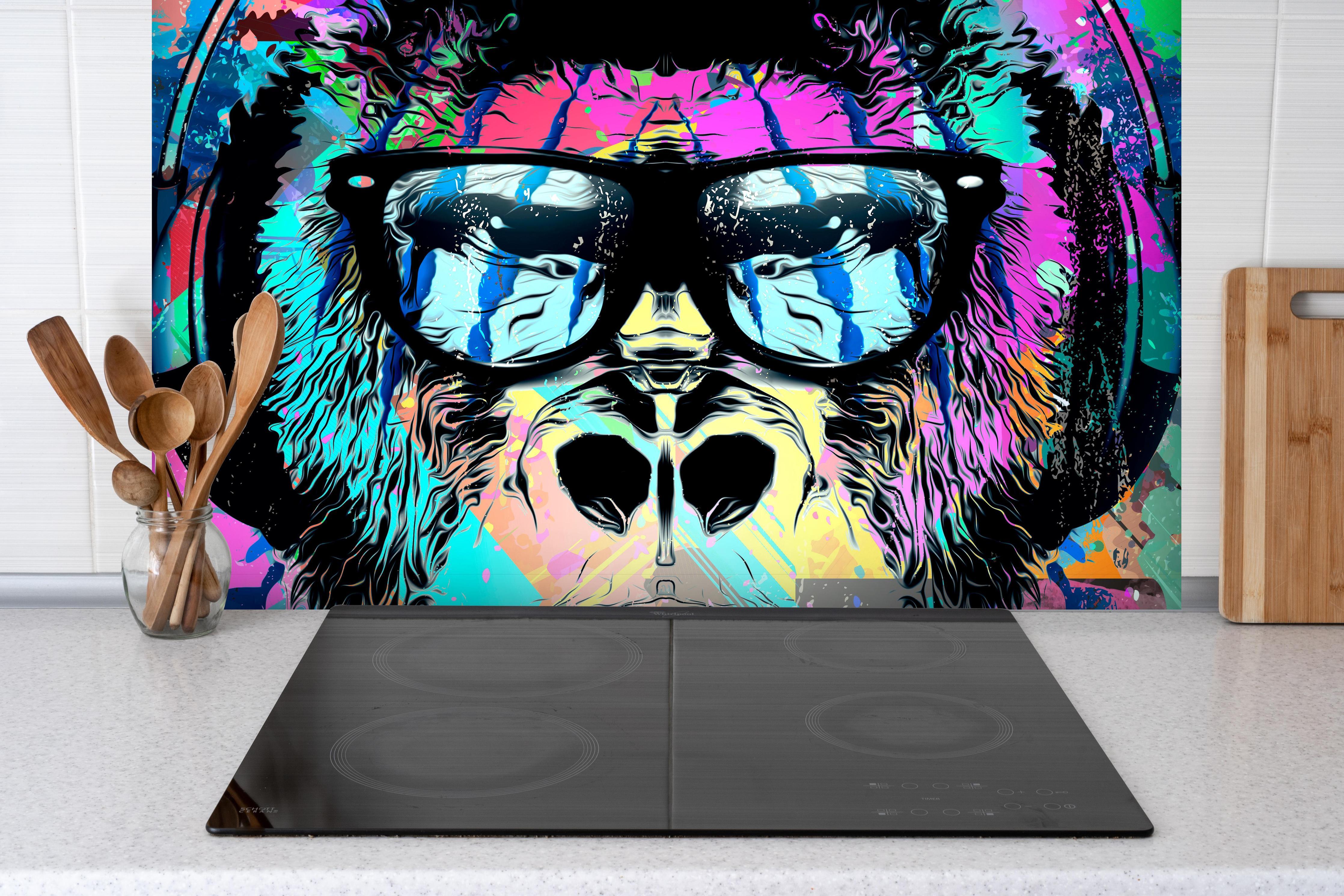 Spritzschutz - Bunter Affenkopf mit Kopfhörer und Brille hinter einem Cerankochfeld zwischen Holz-Kochutensilien