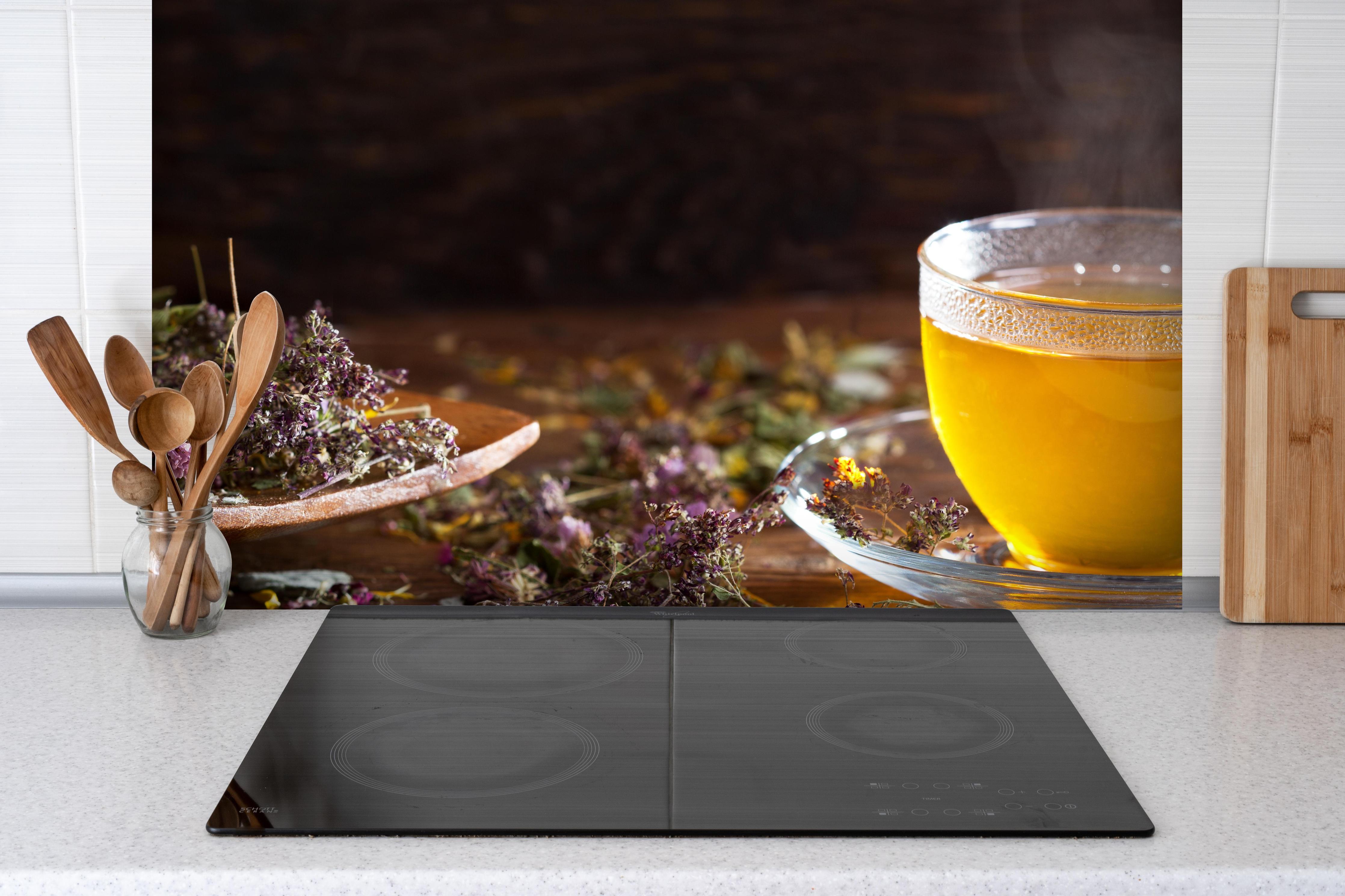 Spritzschutz - Dampfende Tasse Tee mit Kräutern hinter einem Cerankochfeld zwischen Holz-Kochutensilien