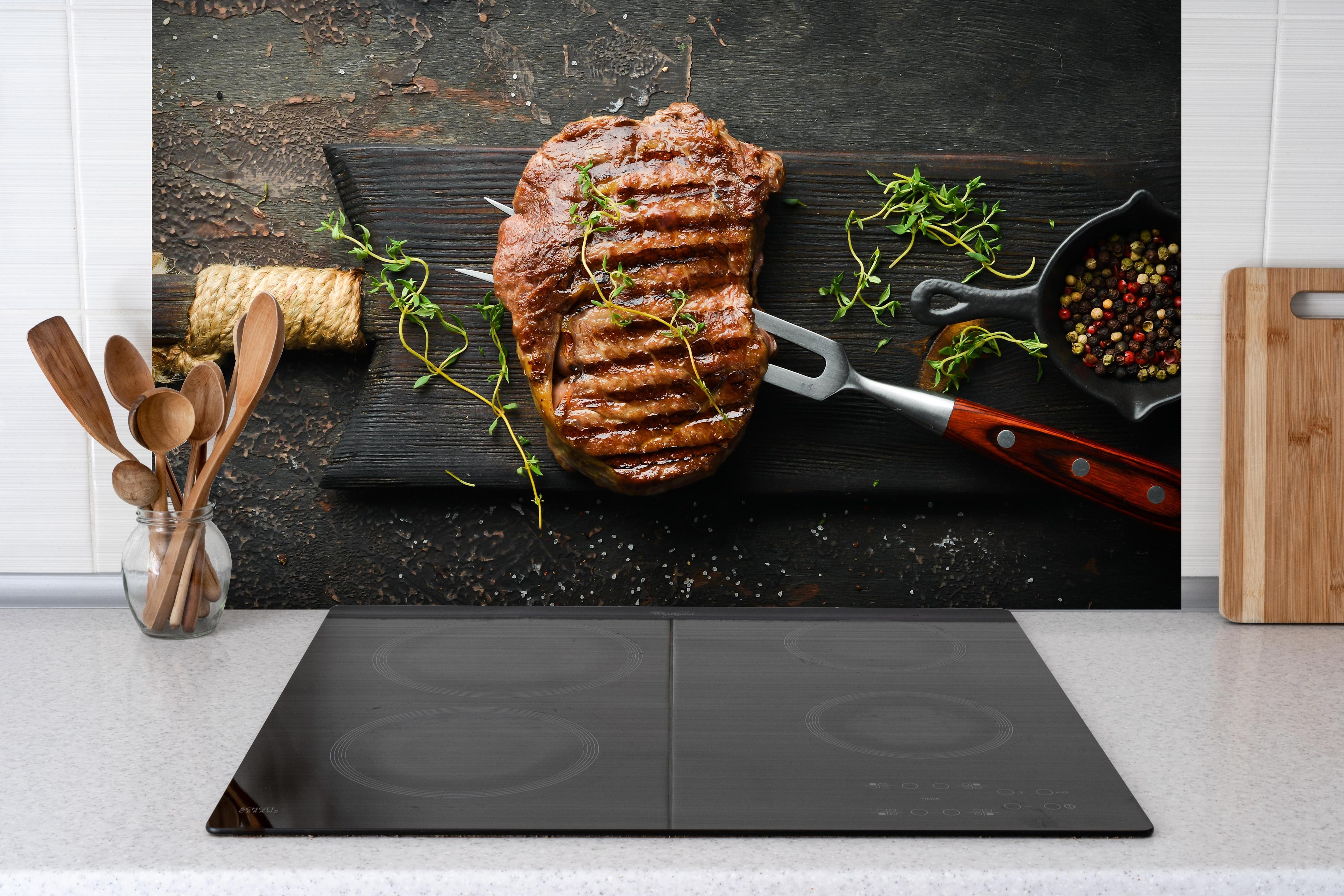 Spritzschutz - Gegrilltes Ribeye auf Holzplatte hinter einem Cerankochfeld zwischen Holz-Kochutensilien