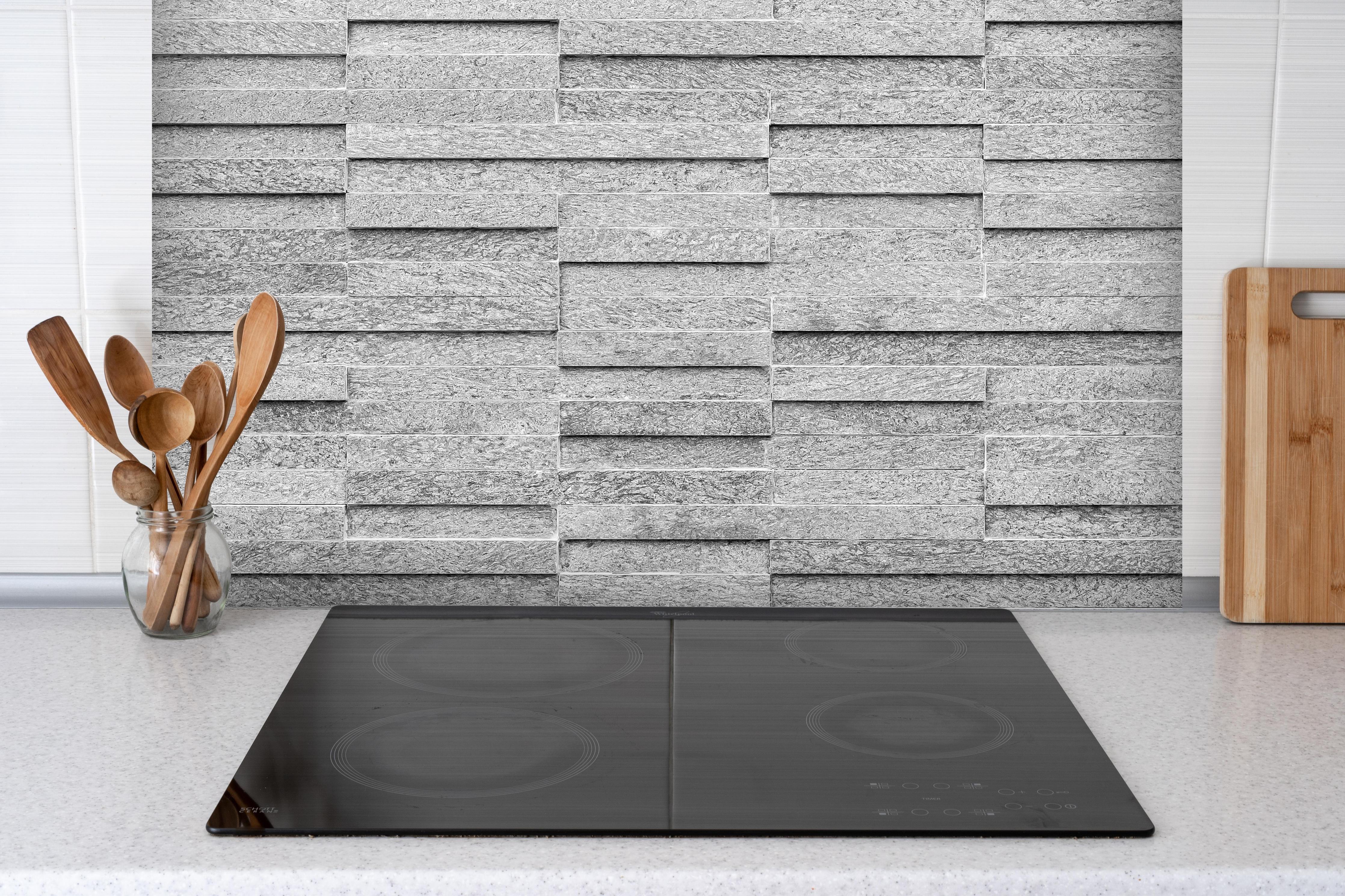 Spritzschutz - Geriffelte gräuliche Backsteinmauer hinter einem Cerankochfeld zwischen Holz-Kochutensilien