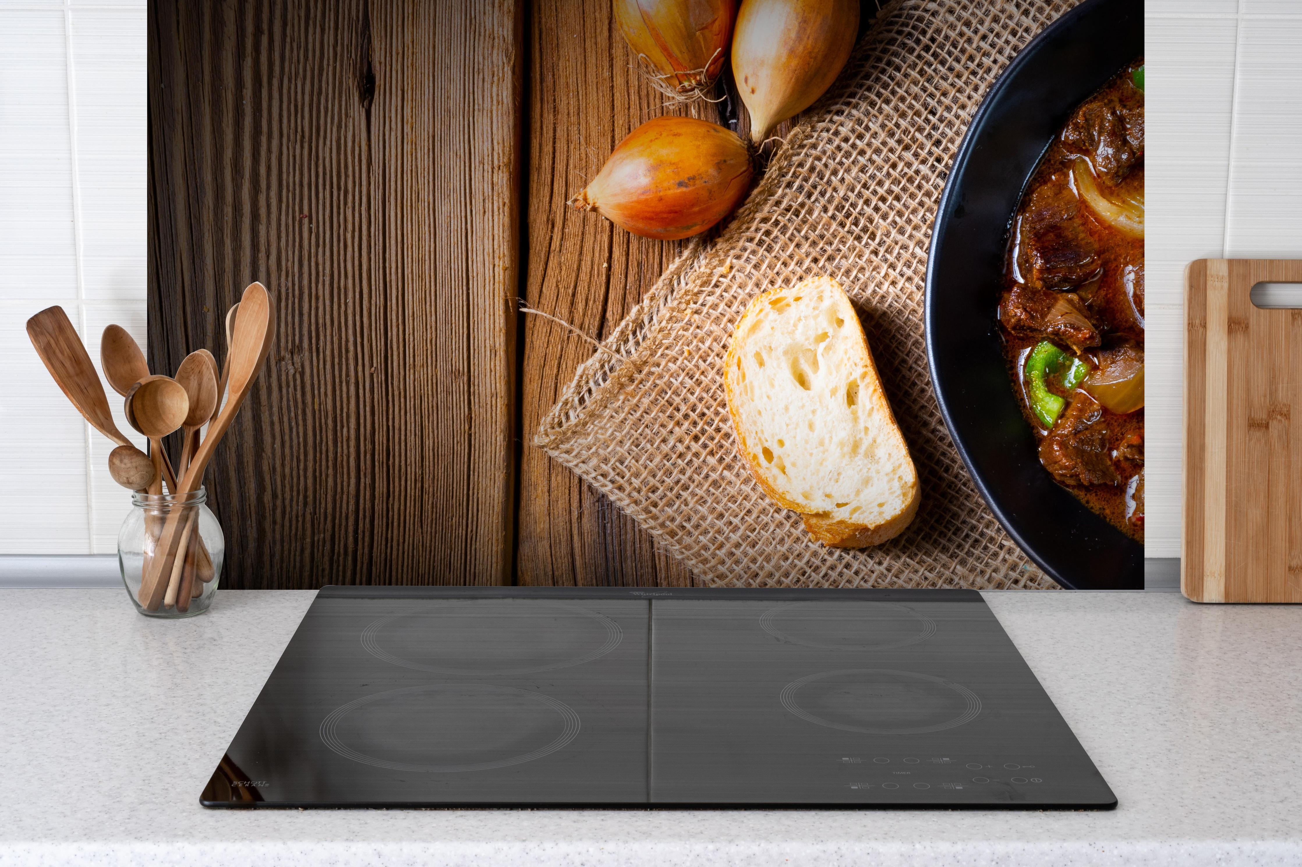 Spritzschutz - Klassisches saftiges Rindergulasch hinter einem Cerankochfeld zwischen Holz-Kochutensilien