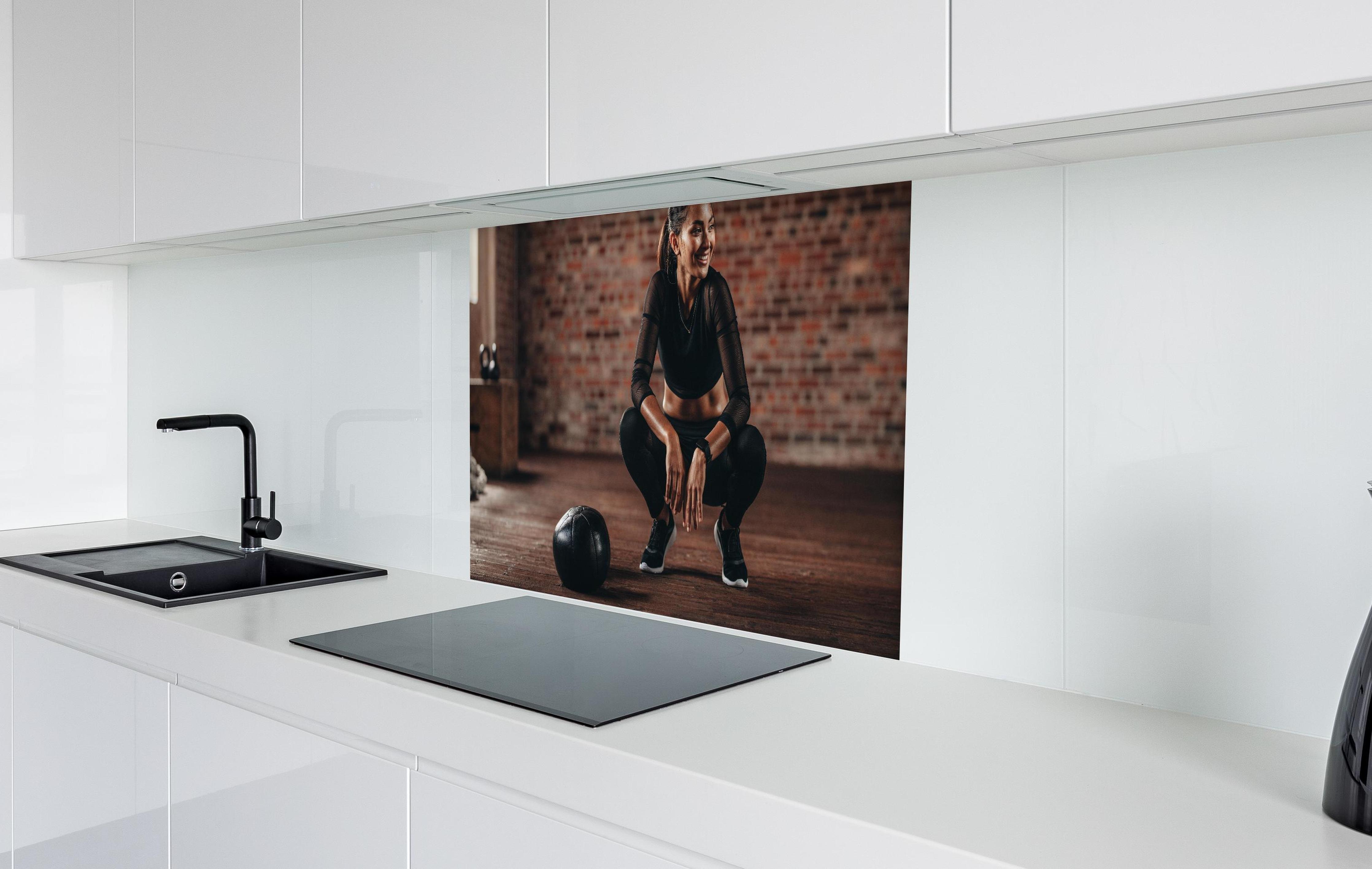 Spritzschutz - Sportlerin in der Hocke in weißer Hochglanz-Küche hinter einem Cerankochfeld