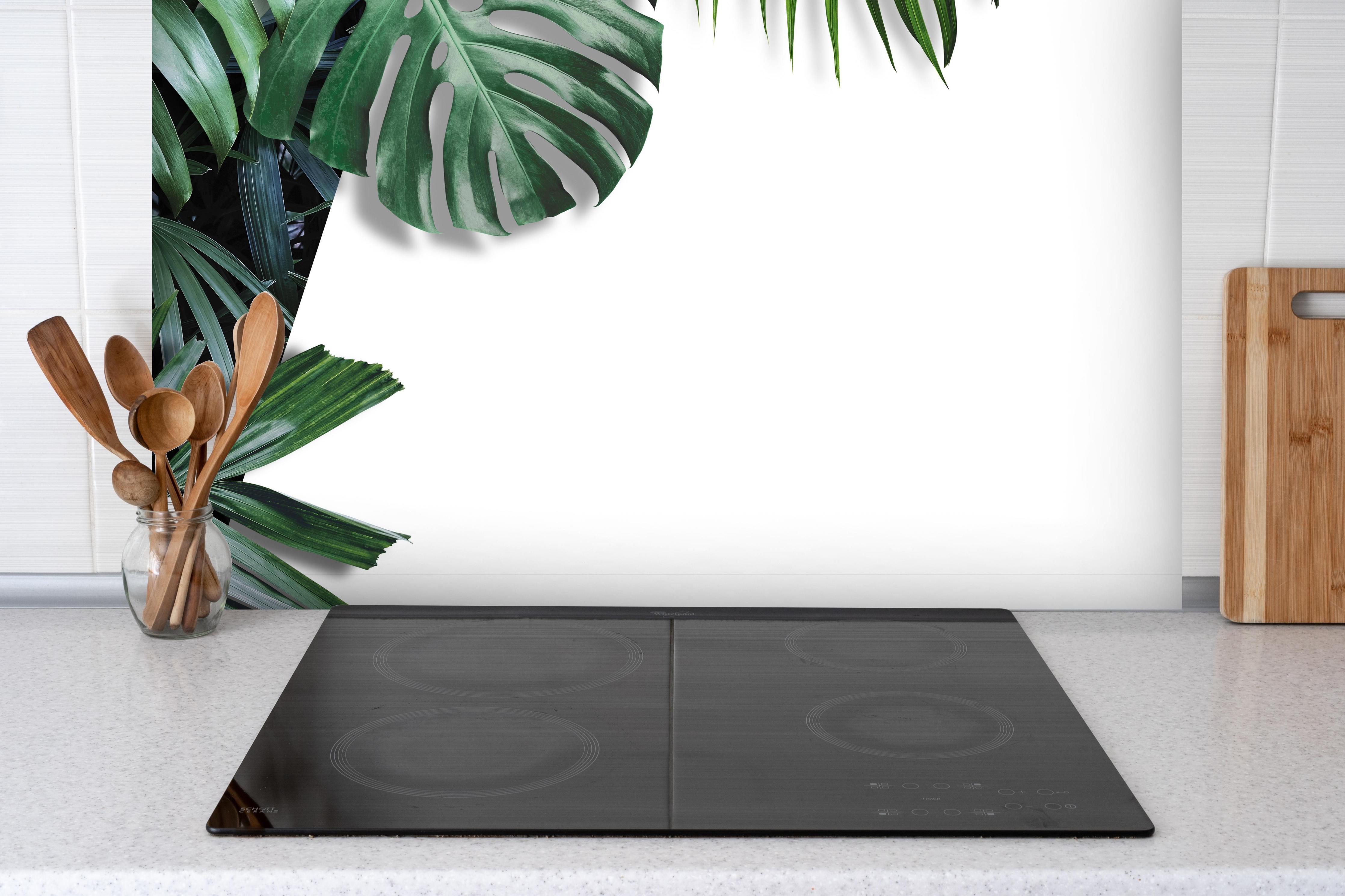 Spritzschutz - Tropische Blätter auf weißem Grund hinter einem Cerankochfeld zwischen Holz-Kochutensilien