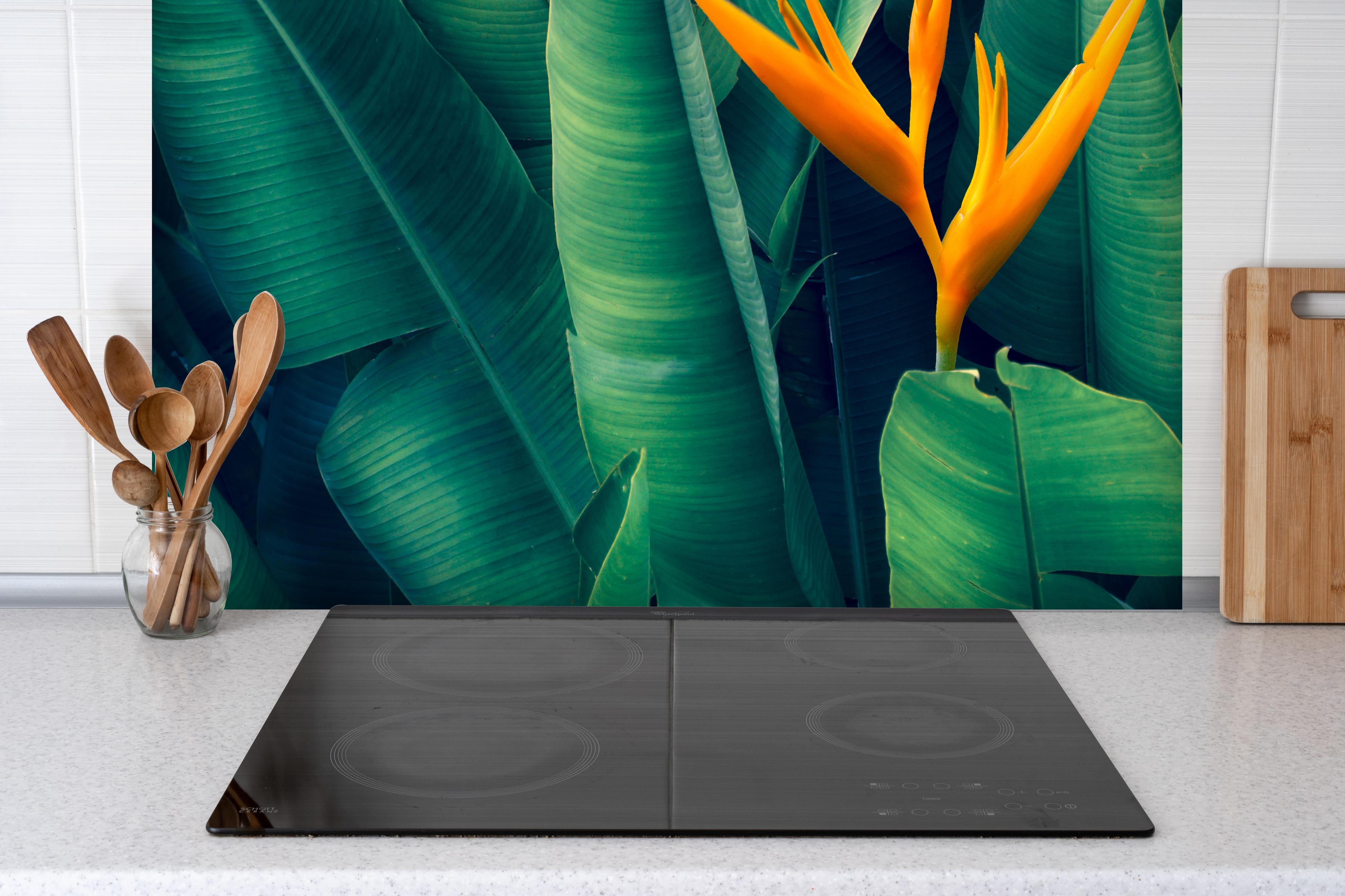 Spritzschutz - Tropische Blätter mit orangefarbiger Blume hinter einem Cerankochfeld zwischen Holz-Kochutensilien