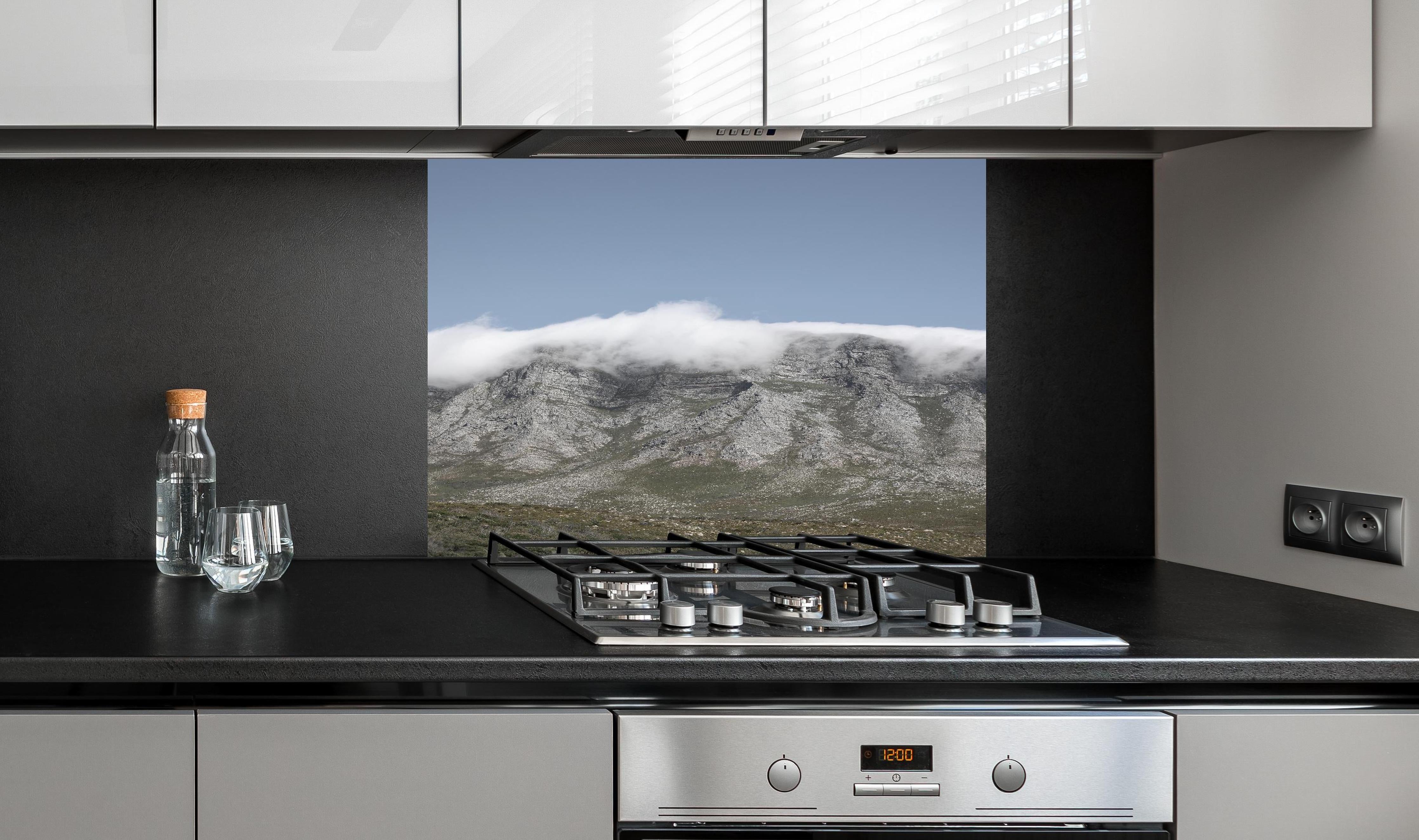 Spritzschutz - Verträumte Berglandschaft mit Wolkenverhang hinter einem Cerankochfeld zwischen Holz-Kochutensilien
