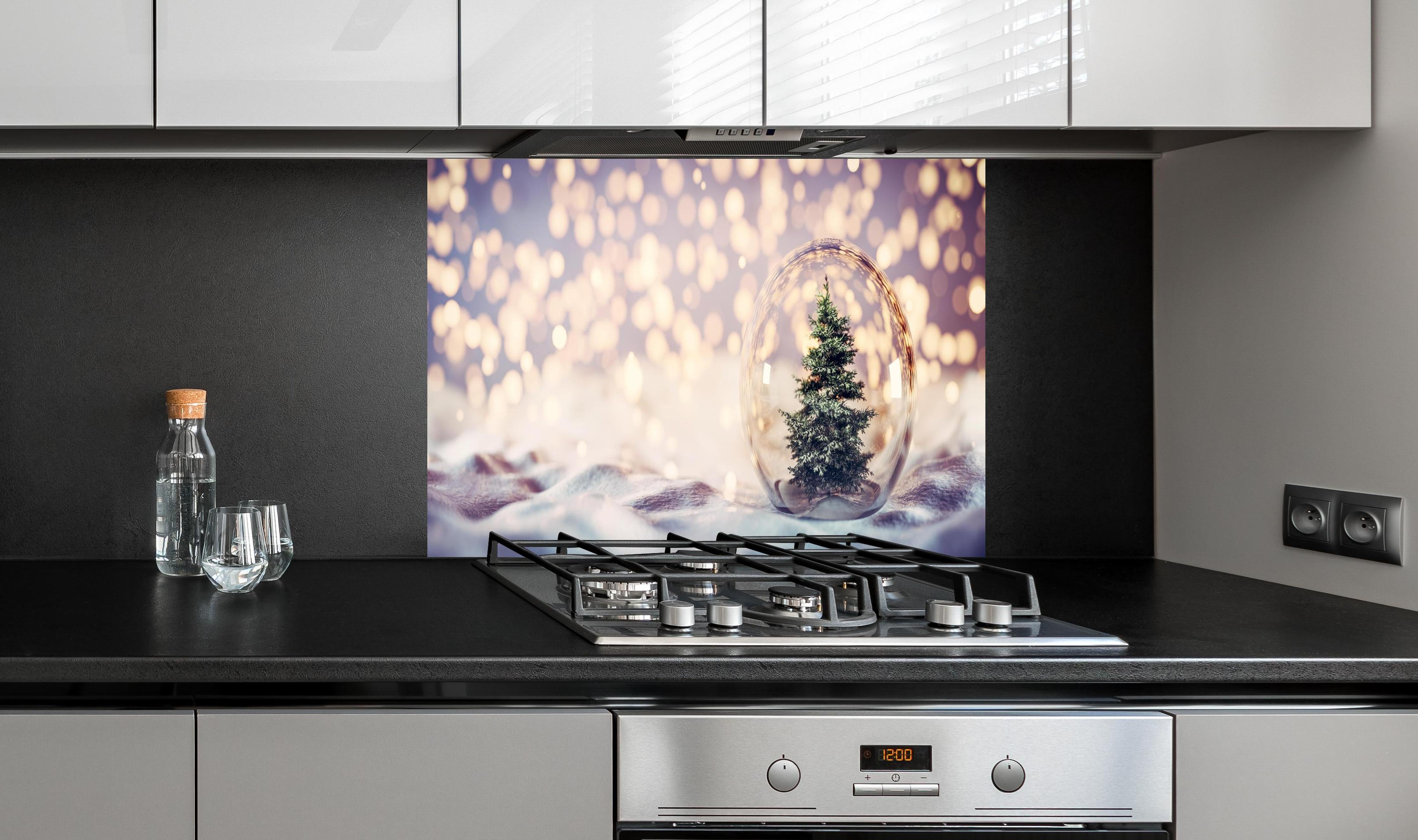 Spritzschutz - Weihnachtsbaum in einer Glaskugel hinter einem Cerankochfeld zwischen Holz-Kochutensilien