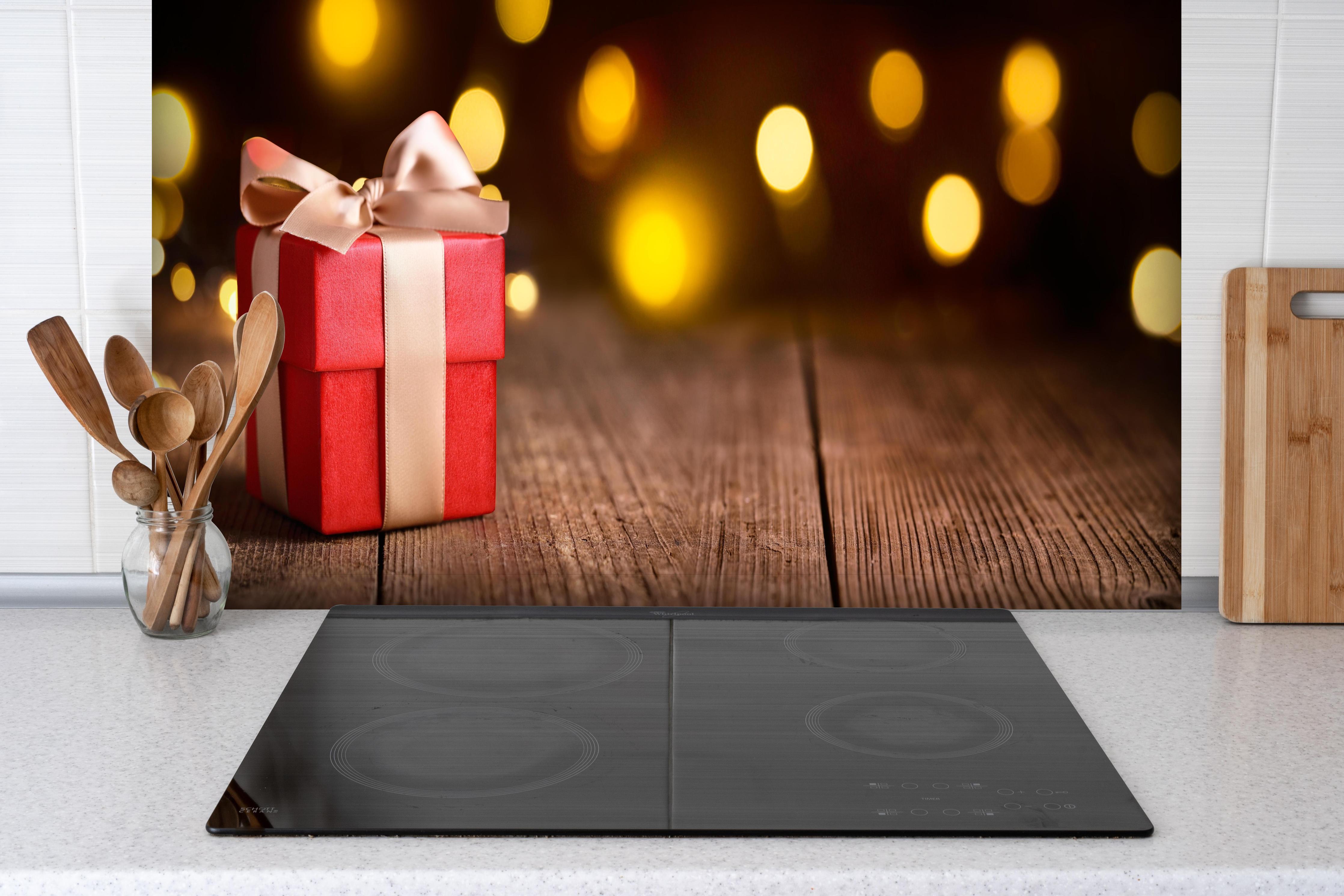 Spritzschutz - Weihnachtsgeschenk auf festlichem Hintergrund hinter einem Cerankochfeld zwischen Holz-Kochutensilien