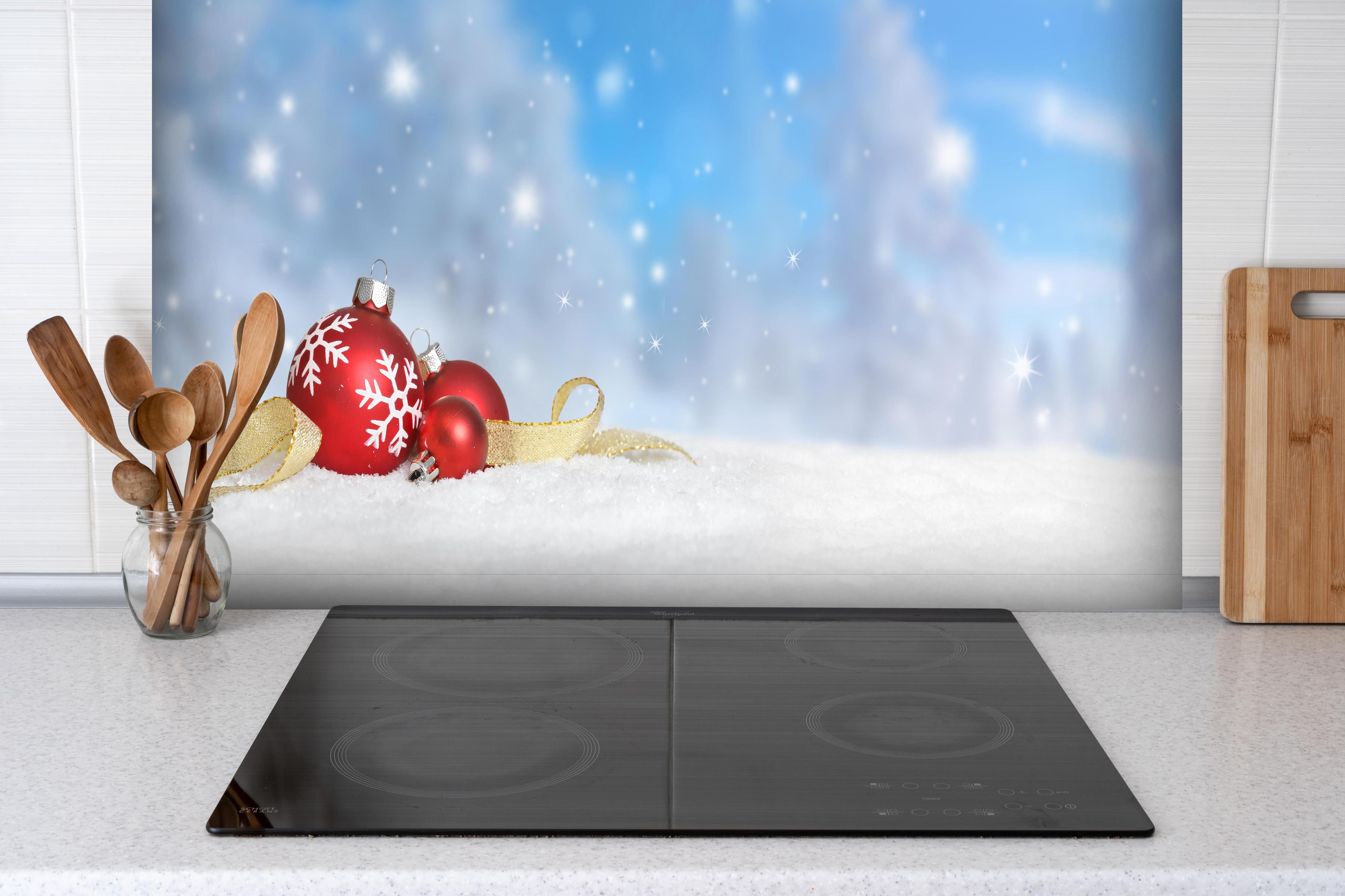 Spritzschutz - Weihnachtskugeln im Schnee hinter einem Cerankochfeld zwischen Holz-Kochutensilien
