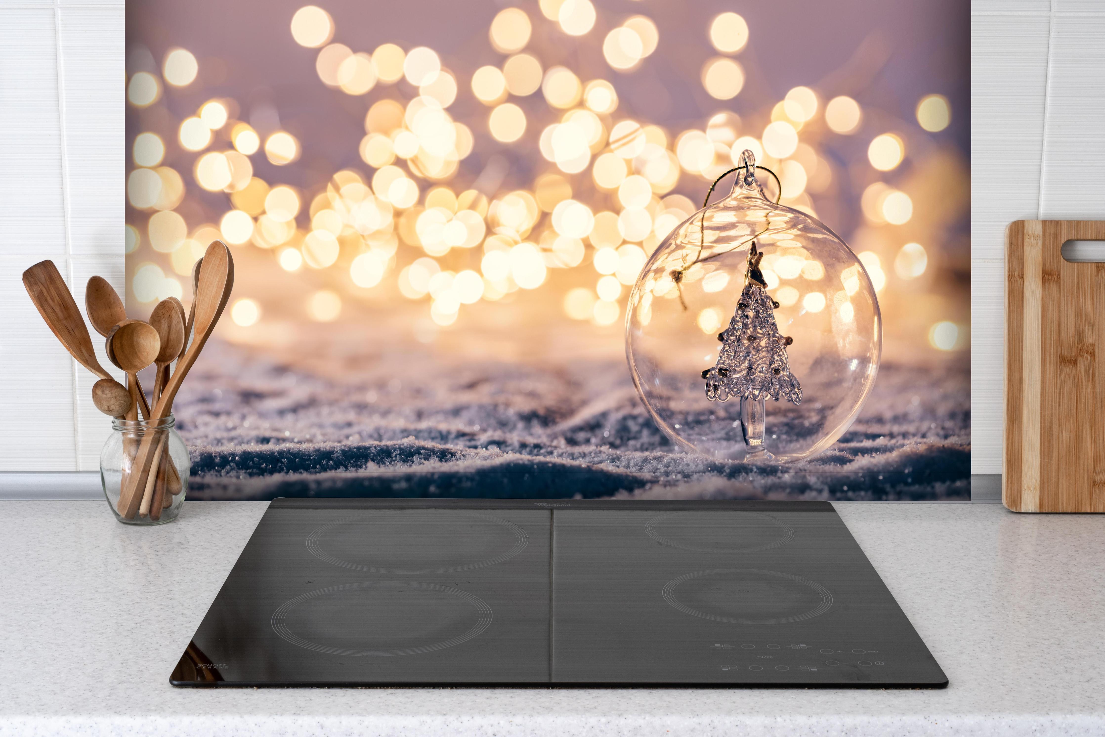 Spritzschutz - Winterliche Weihnachtskugel mit Baum-Motiv hinter einem Cerankochfeld zwischen Holz-Kochutensilien