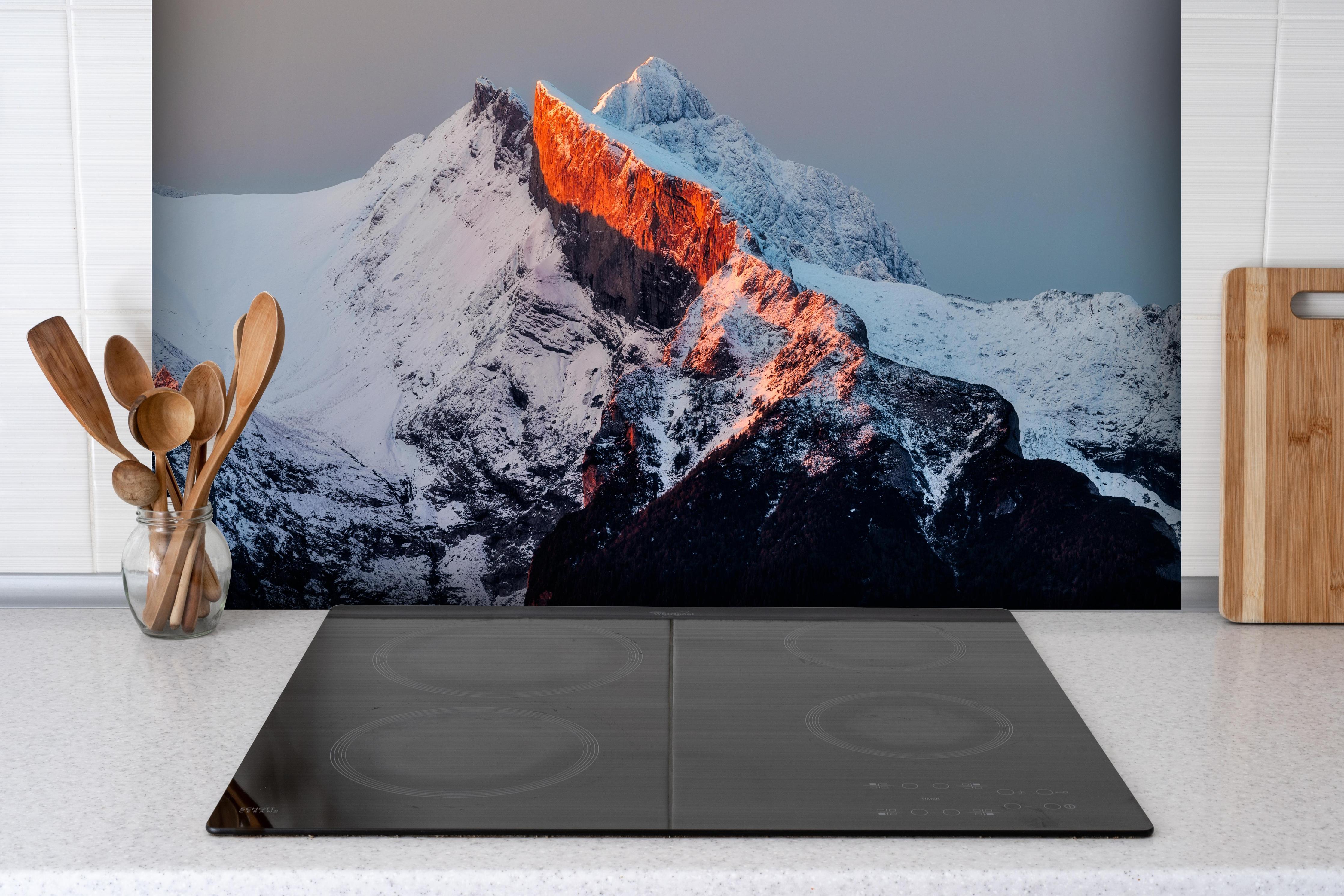 Spritzschutz - Winterlicher Sonnenaufgang über schneebedeckten Gipfeln hinter einem Cerankochfeld zwischen Holz-Kochutensilien