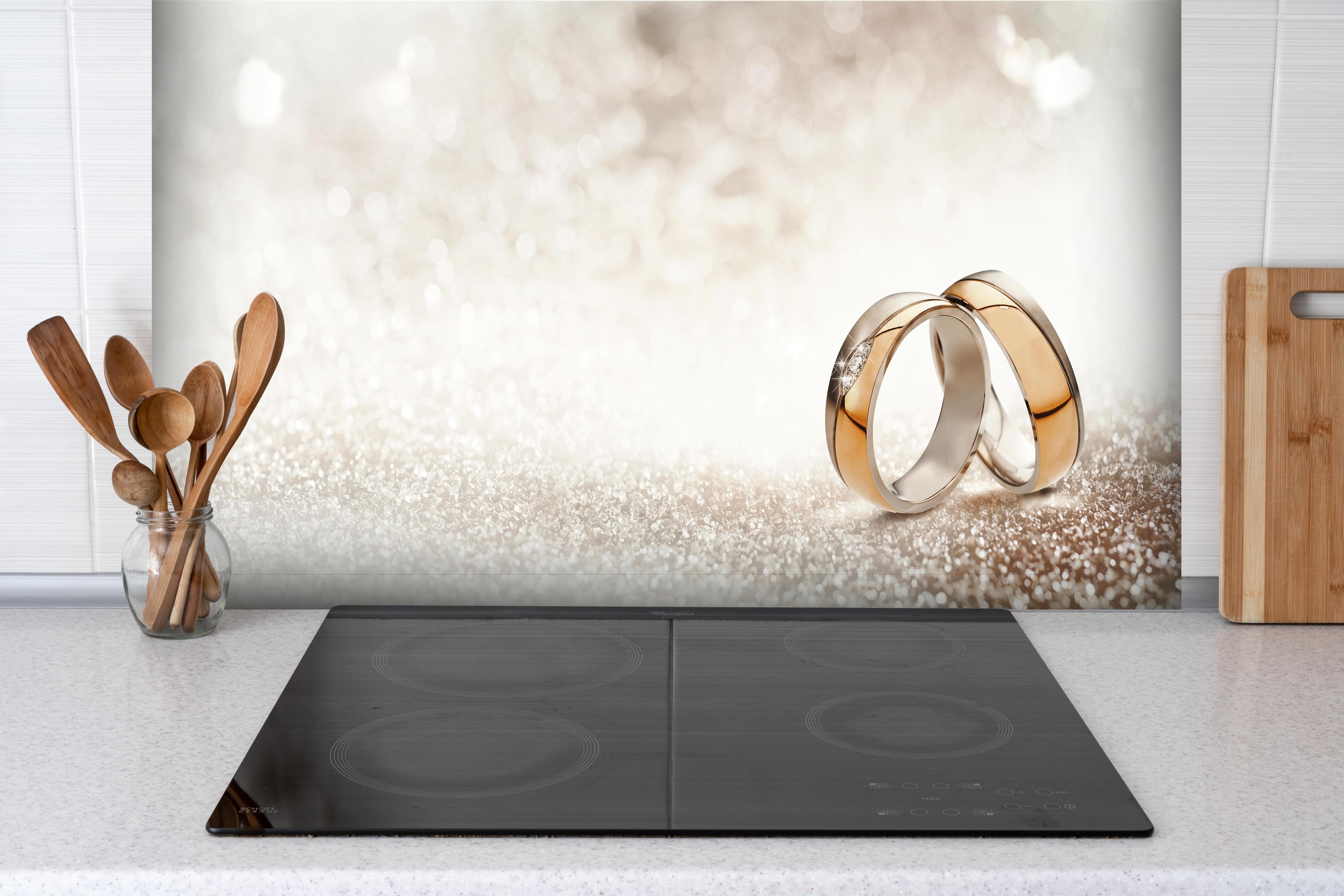 Spritzschutz - Zwei goldene Eheringe mit Glitter Hintergrund hinter einem Cerankochfeld zwischen Holz-Kochutensilien