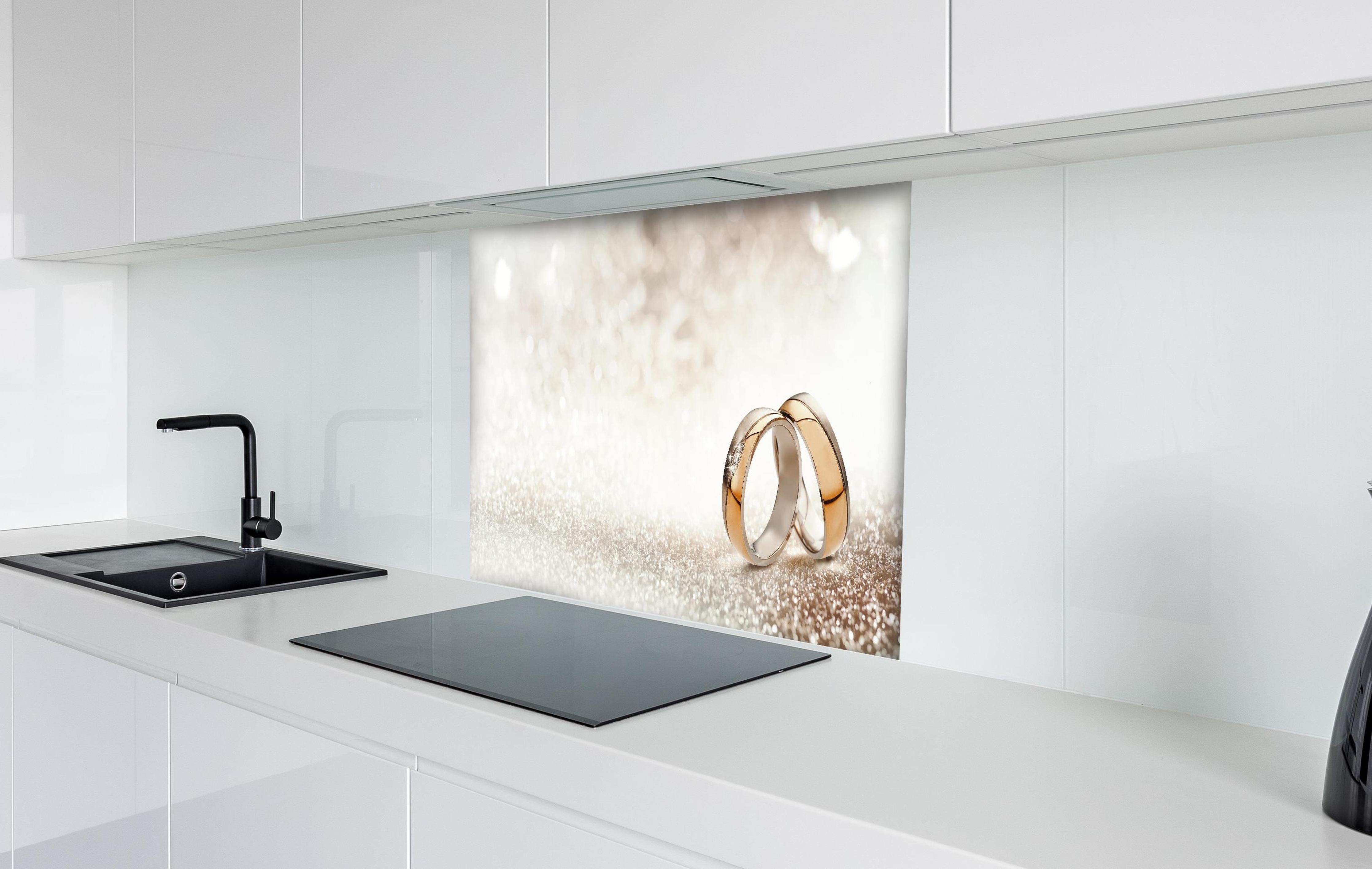 Spritzschutz - Zwei goldene Eheringe mit Glitter Hintergrund hinter einem Cerankochfeld zwischen Holz-Kochutensilien