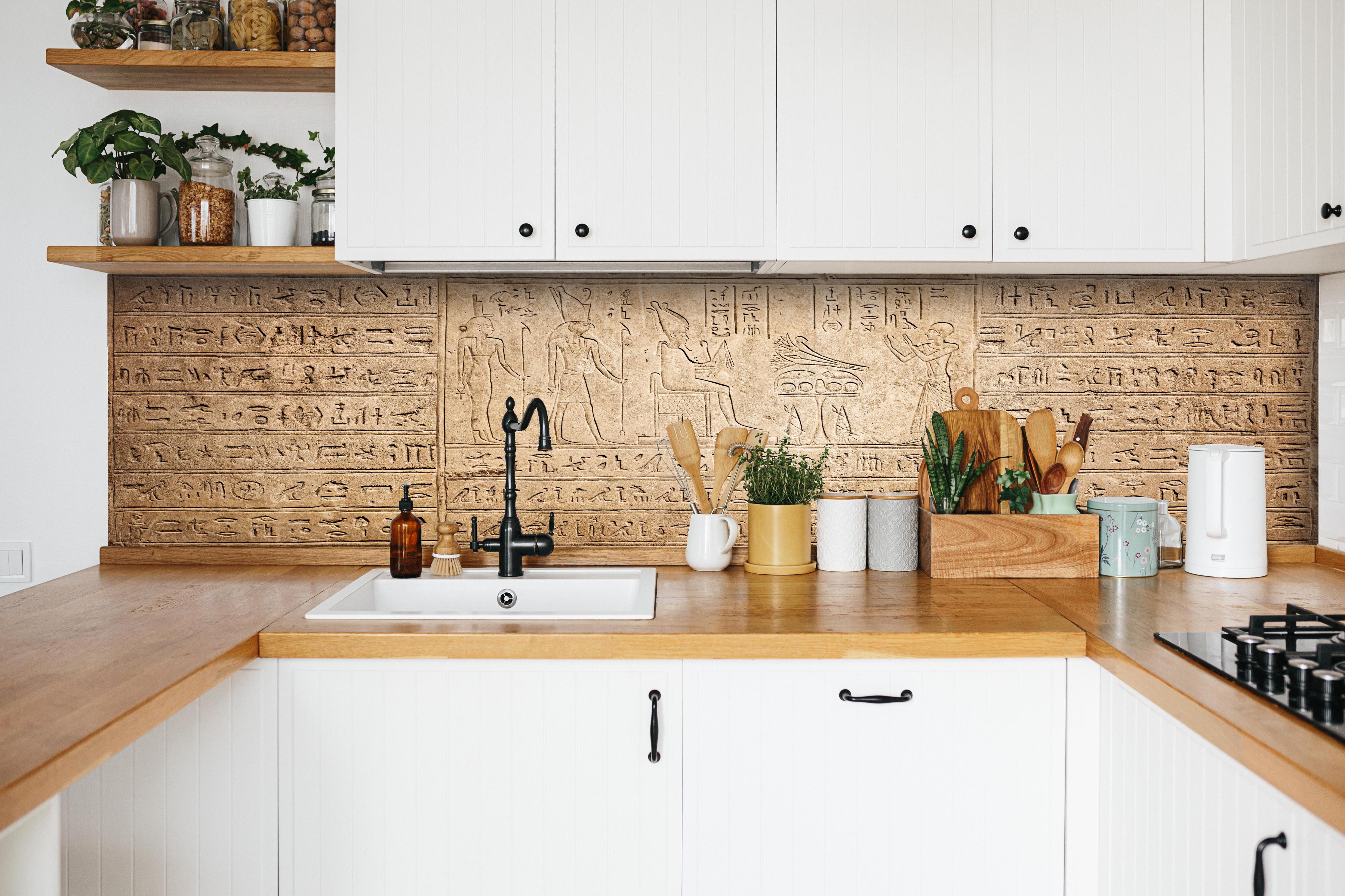 Küche - Ägyptische Hieroglyphen auf Sandstein hinter weißen Hochglanz-Küchenregalen und schwarzem Wasserhahn
