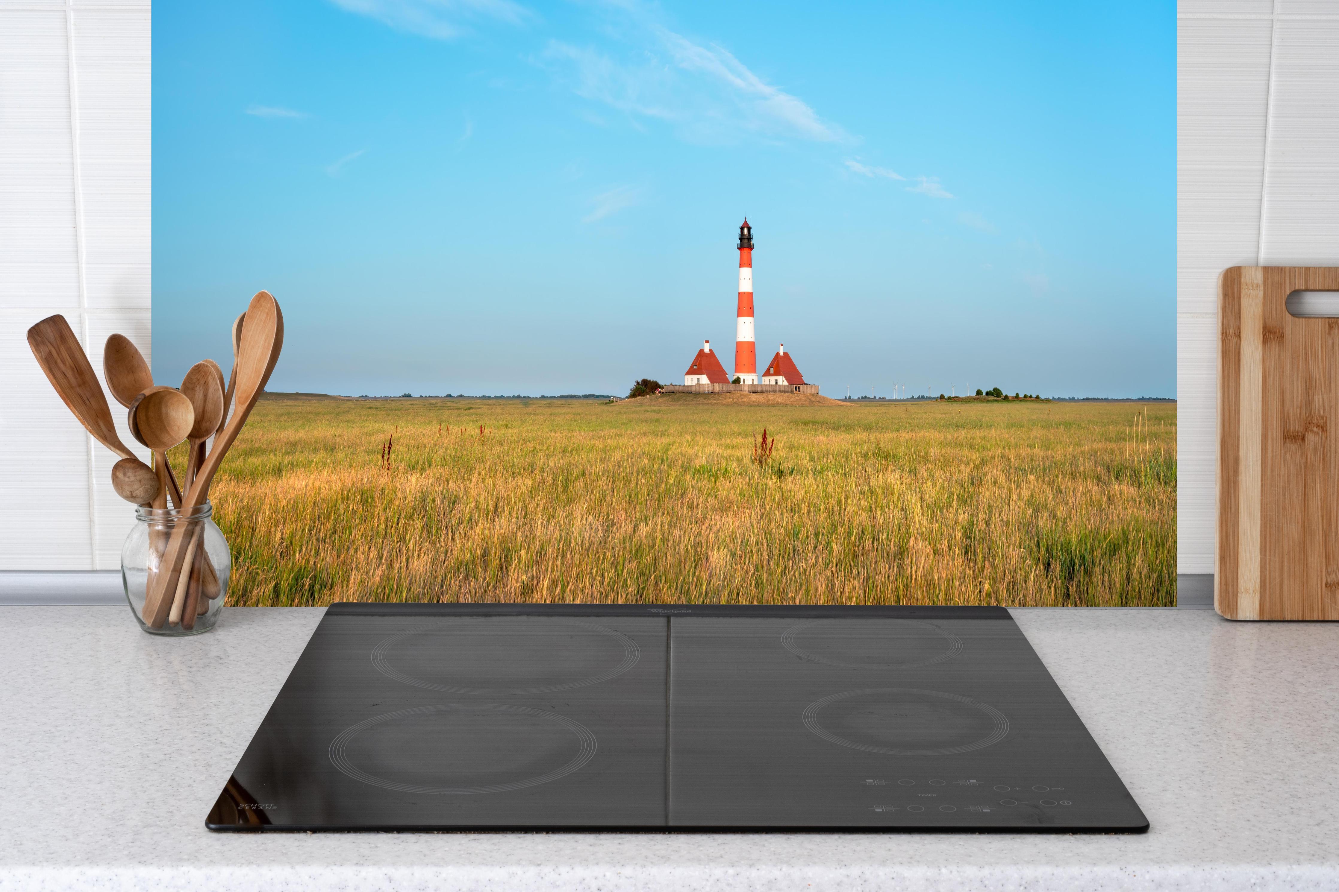 Küche - Ein beeindruckender Leuchtturm in der weitläufigen norddeutschen Landschaft unter blauem Himmel. hinter weißen Hochglanz-Küchenregalen und schwarzem Wasserhahn