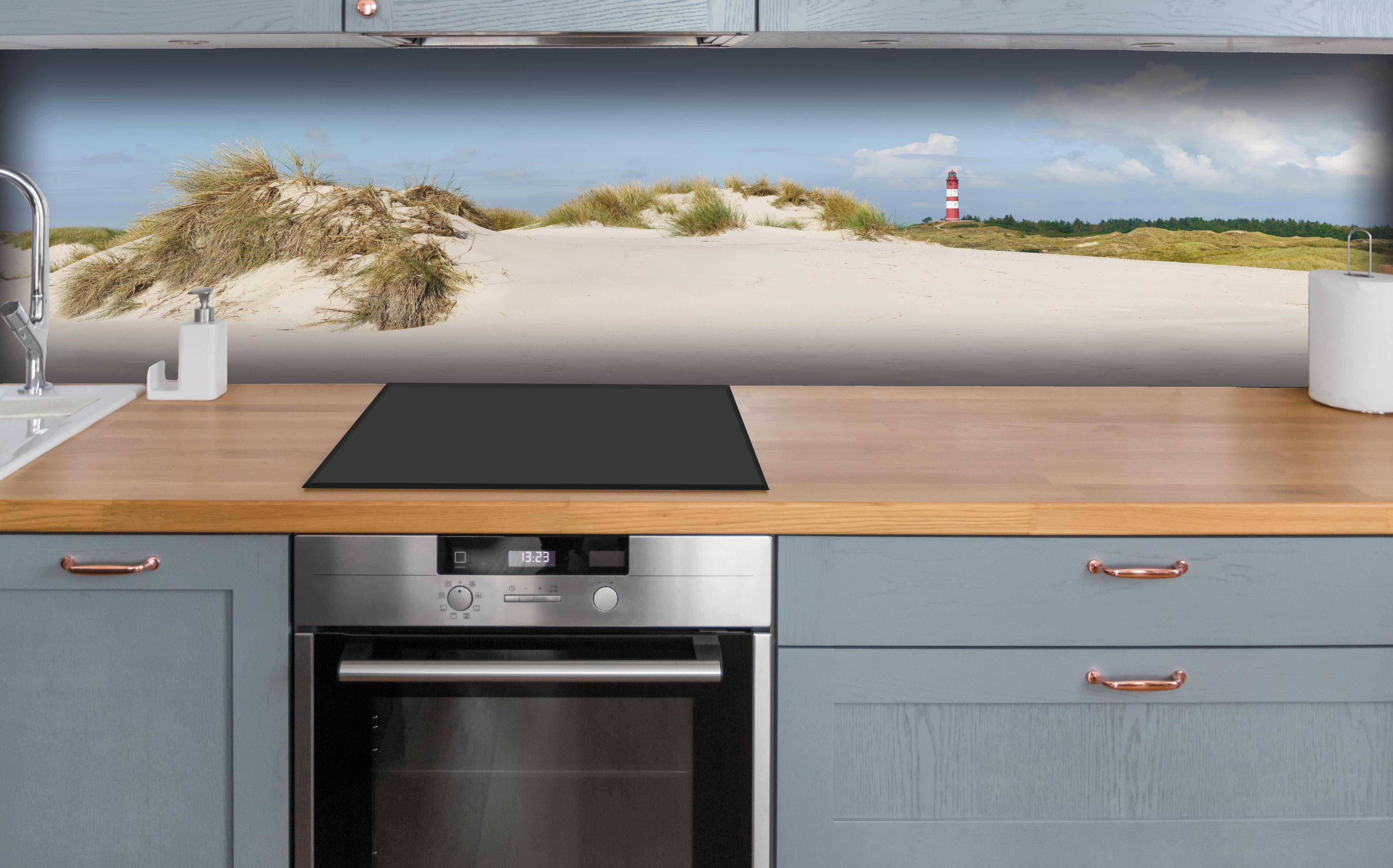 Küche - Ein traumhafter Blick auf die weiße Sanddüne und den roten Leuchtturm am Meer hinter weißen Hochglanz-Küchenregalen und schwarzem Wasserhahn