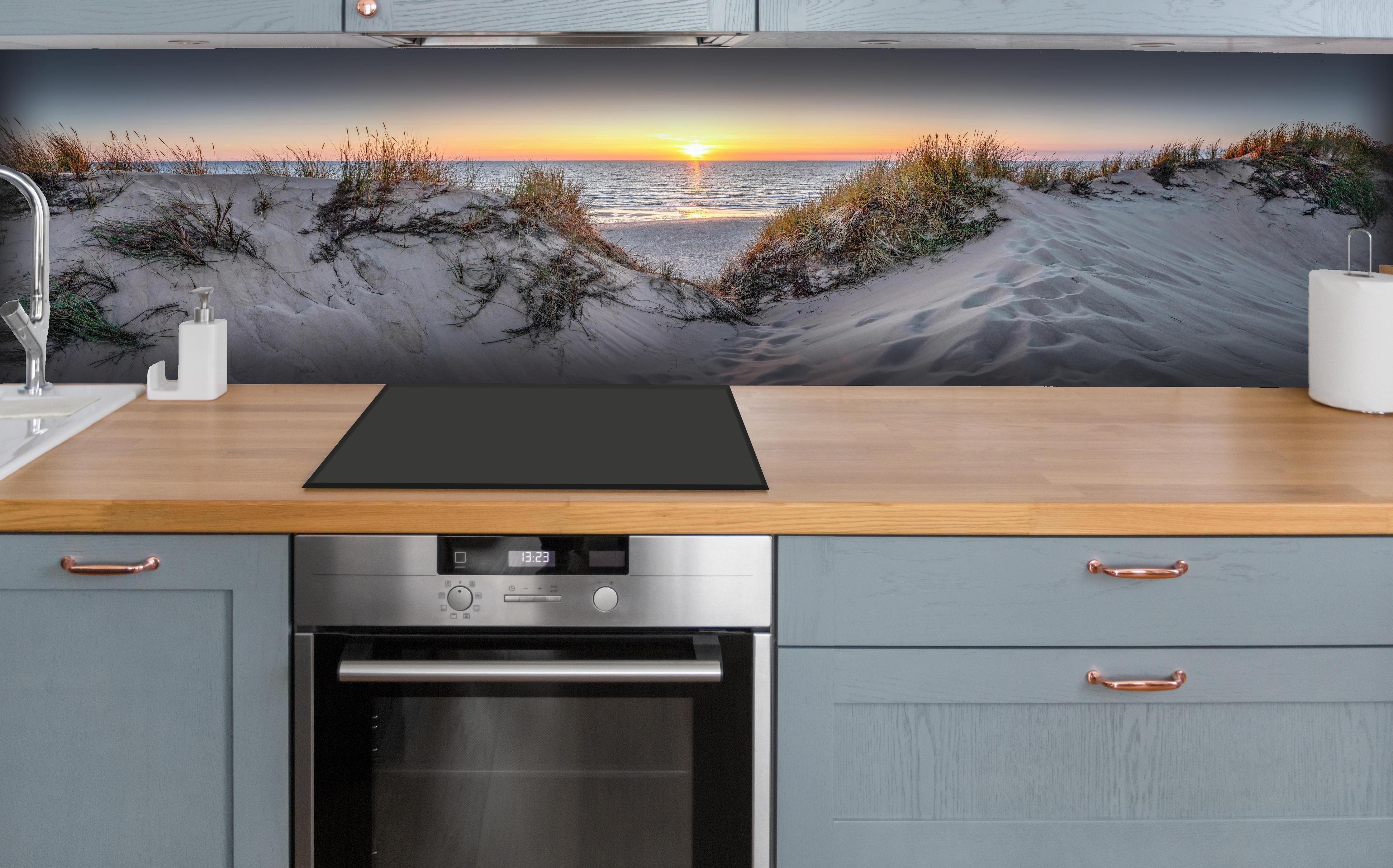Küche - Faszinierender Sonnenuntergang über einer Sanddüne am Meer hinter weißen Hochglanz-Küchenregalen und schwarzem Wasserhahn
